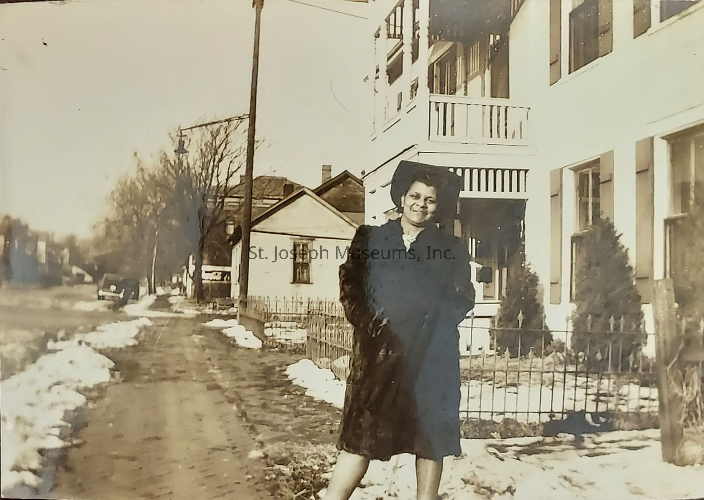 In this vintage black-and-white photograph, a woman stands confidently near the fence of a large residential building on a sidewalk lined with patches of snow. Dressed in a warm coat and a stylish hat, she smiles softly, exuding a sense of elegance and composure. On the right side of the image, the building she leans against features multiple stories and balconies, suggesting it is a substantial home or perhaps an apartment complex. In the background, smaller houses and leafless trees line the street, alongside a parked car further down the road, hinting at a quaint neighborhood setting. The overall atmosphere is calm and nostalgic, capturing a moment from a bygone era, detailed with the seasonal evidence of winter and early 20th-century fashion.