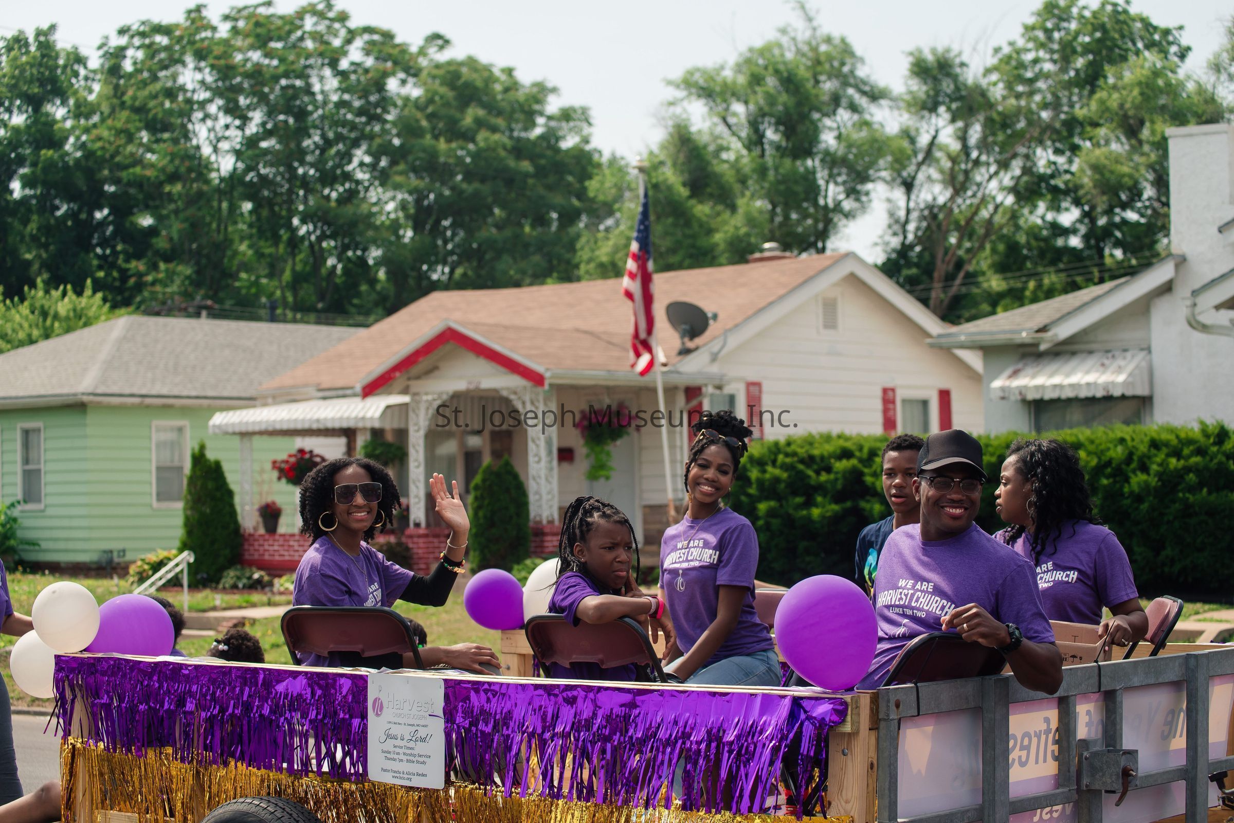 The image shows a group of people participating in a parade, riding on a float decorated with purple and gold streamers. They are seated and waving, appearing to enjoy the festive atmosphere. Some are wearing matching purple T-shirts that read "We Are Harvest Church" with joy and togetherness evident in their smiles. The float is also adorned with purple and white balloons, enhancing the cheerful and celebratory scene. Behind them, there's a charming neighborhood with small houses and green trees. An American flag stands prominently in the background, contributing to the festive, community-centered vibe of the parade.