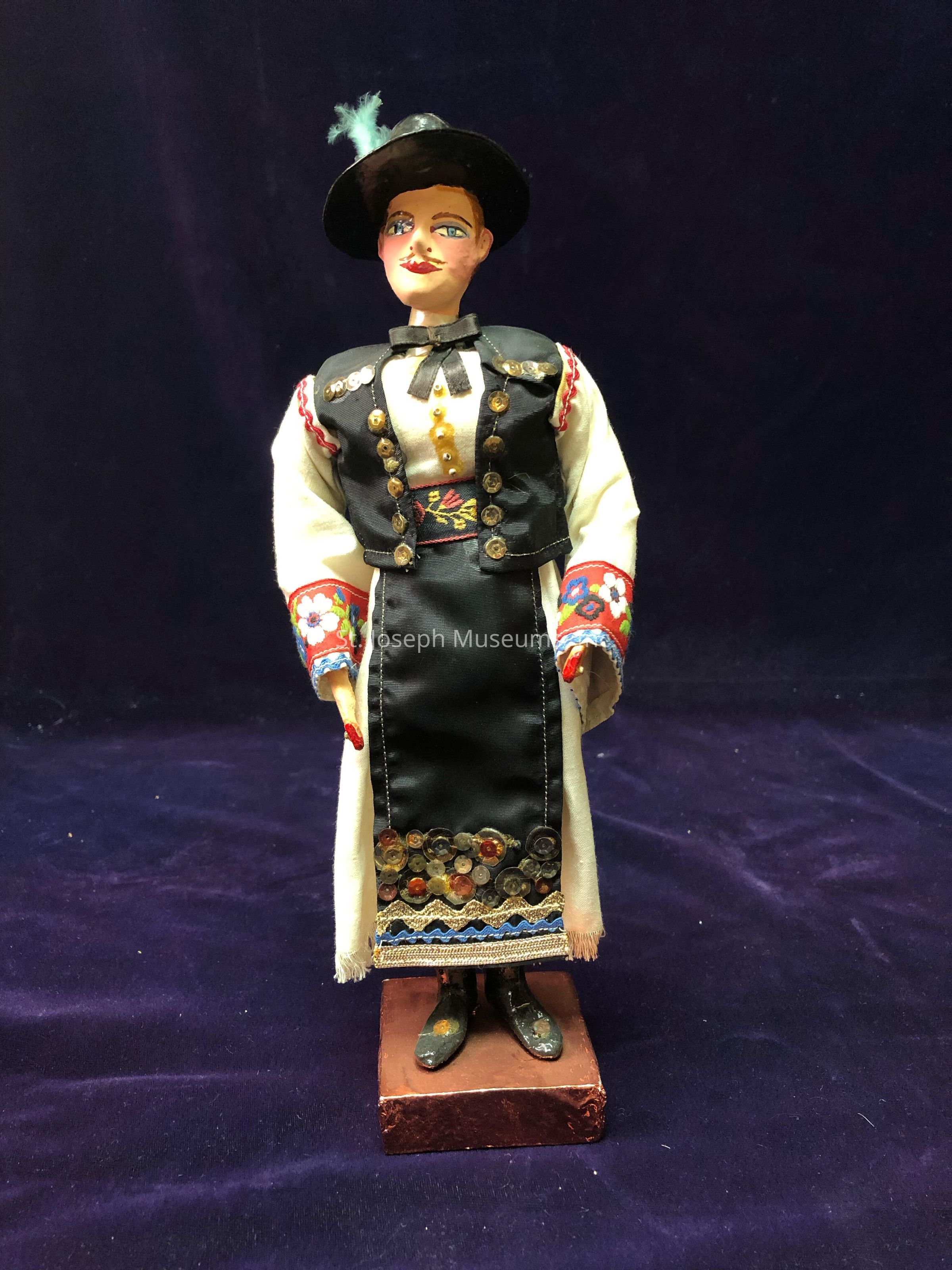 The image shows a handcrafted doll standing on a square base against a dark blue background, wearing a traditional-style outfit. The attire consists of a black vest adorned with golden buttons over a white shirt, featuring a decorative black skirt that reaches below the knees. The sleeves of the shirt are adorned with vibrant, intricate floral embroidery in red, blue, and white. The doll's accessories include a small black hat with a feather and black shoes. Its painted facial features include blue eyes and red lips, complemented by delicately painted cheeks. The overall composition presents an impression of tradition and cultural richness, highlighted by the detailed design work and choice of colors. The fabric and materials used give the doll a textured, artful appearance, embodying a sense of craftsmanship and heritage. This figurine stands gracefully on a dark purple fabric surface, distinctively positioned at the center of the image, where its elements and characteristics draw attention to its cultural and artistic significance.