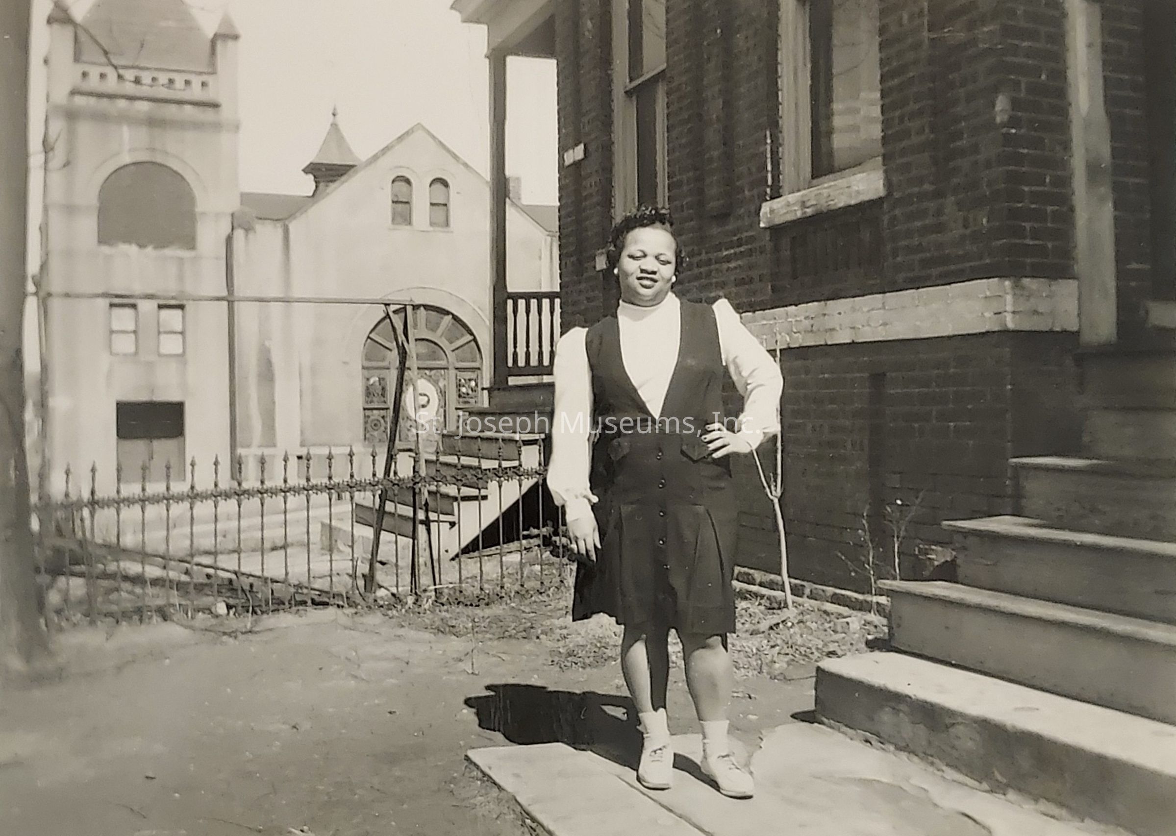 The black and white photo depicts a woman standing confidently with one hand on her hip in front of a brick building, near a flight of steps and a metal railing. She is dressed in a dark button-up dress over a white blouse, and wears white socks with light-colored shoes. In the background, a large, ornate building with arched windows and a small spire is visible, suggesting an urban setting. The scene is lit by natural daylight, casting soft shadows on the ground, adding to the nostalgic and historical ambiance of the image.