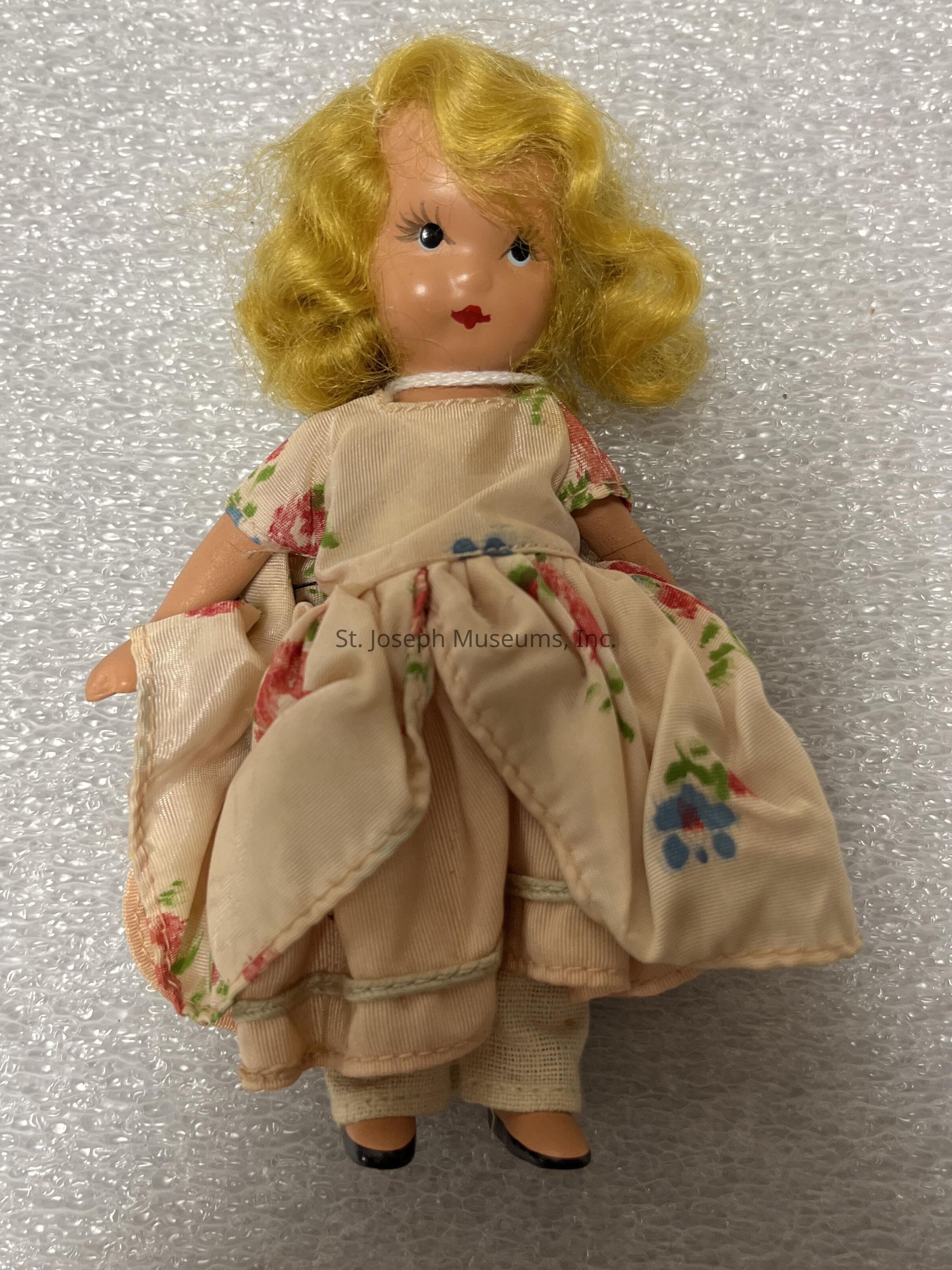 The image displays a vintage doll with curly, blonde hair and a delicate expression, featuring hand-painted facial details like wide eyes and red lips. She is wearing a cream-colored dress adorned with floral patterns in shades of red and blue, adding a touch of colorful elegance. Her attire includes a full skirt, giving the doll a classic, old-fashioned look. The doll's arms are slightly bent, and she wears black shoes peeking out from beneath her dress. The background consists of bubble wrap, emphasizing the preservation and care taken for this collectible item, suggesting it might hold sentimental or historical value.