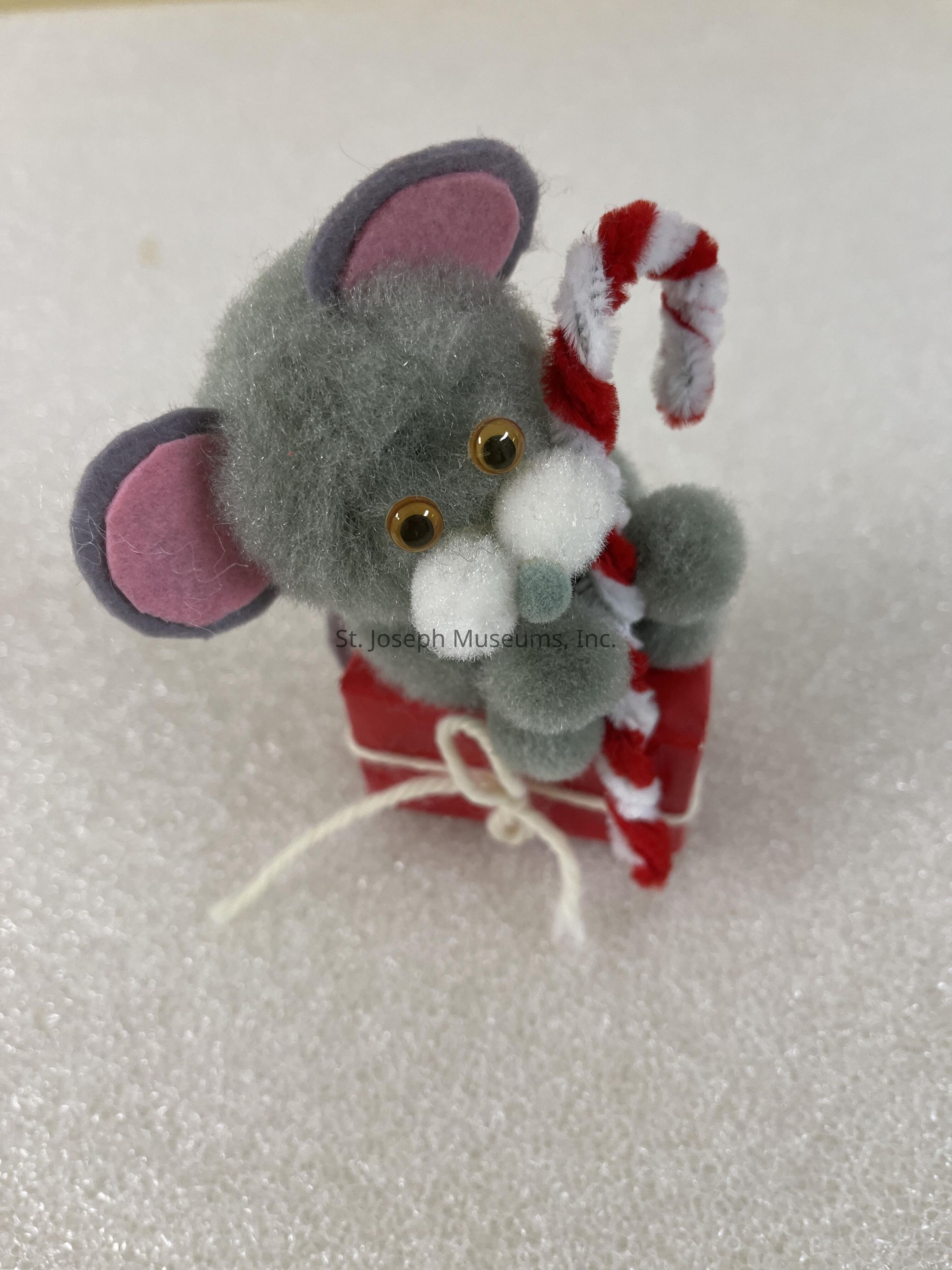 The image depicts an adorable, small, plush toy mouse with gray fur, pink inner ears, and round golden eyes, holding a red and white striped candy cane. This cute figure, crafted with fuzzy material, is sitting on a tiny red box tied with a white string, resembling a present, against a soft, white textured background. The combination of the mouse's whimsical design and the festive elements evokes a sense of holiday cheer and playful charm.