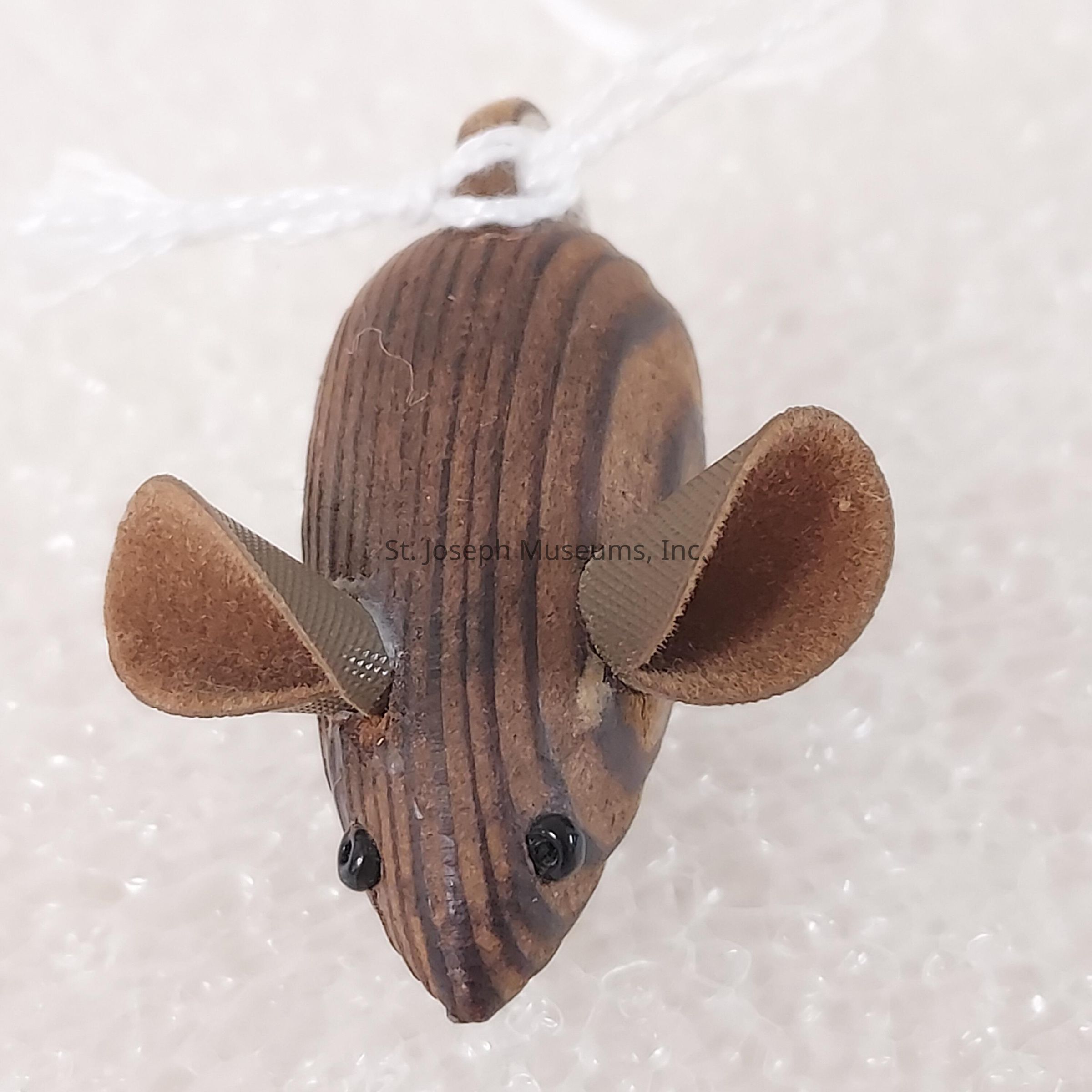 The image depicts a small, handcrafted mouse figurine, skillfully made from wood with a smooth, polished appearance. The body showcases natural wood grain patterns, contributing to its rustic charm. It features a pair of large, rounded ears made from a soft, felt-like material that gives the figurine a playful and whimsical look, and two tiny, shiny black beads represent the eyes, adding detail and character. The nose is pointed, enhancing its mouse-like appearance, while the texture is further enriched by subtle carving lines running along the length of the body. A white string loop is attached at the top, suggesting that it could be used as a decoration or ornament. The mouse is set against a soft, textured background that resembles porous material, which contrasts gently with the figure's darker tones, ensuring that the small details and craftsmanship are visibly appreciated. Overall, the object is a charming and creative representation of a mouse, demonstrating fine craftsmanship and attention to detail, making it a delightful decorative piece.