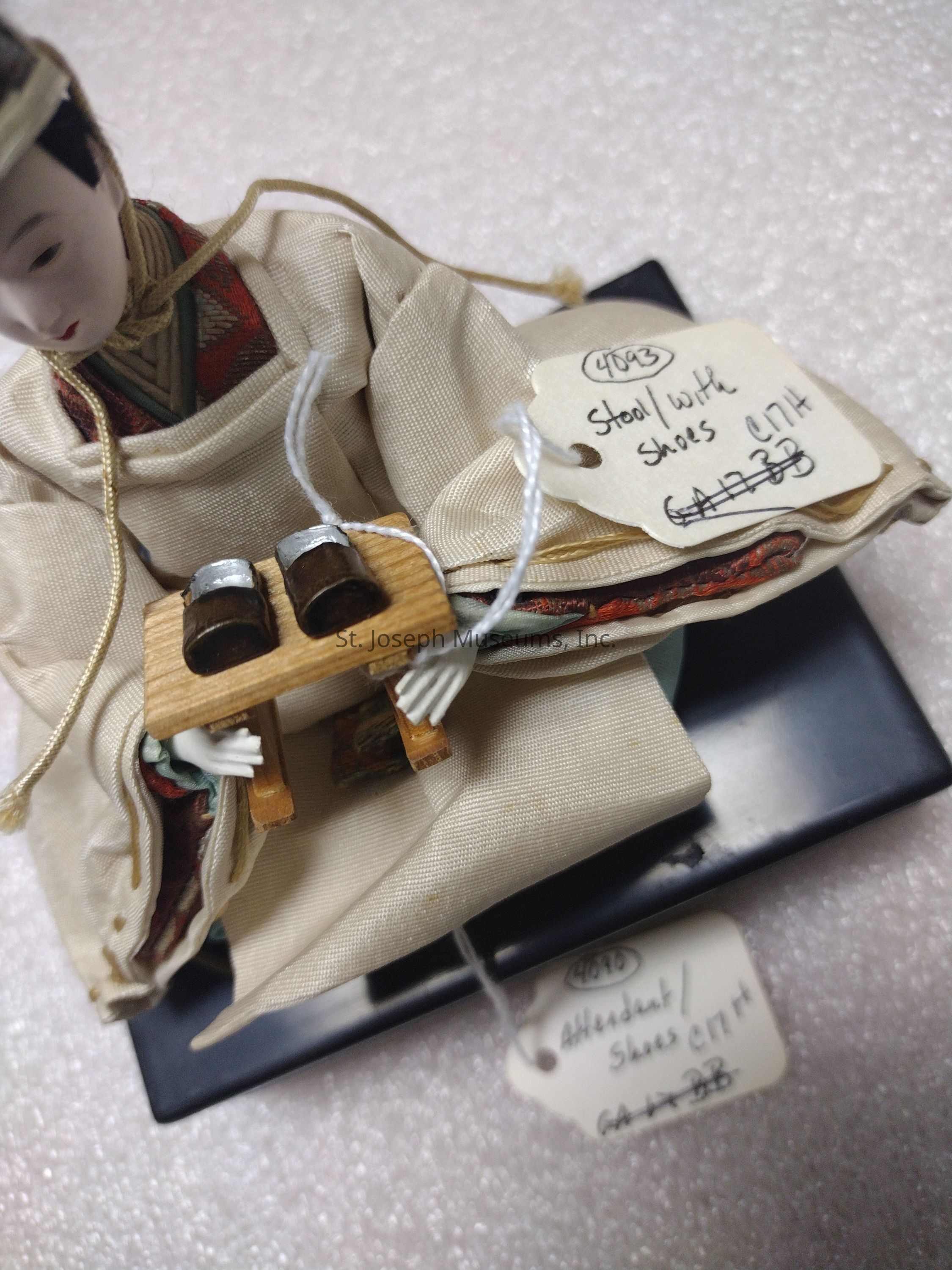 The image features a detailed figurine dressed in traditional attire, seated and holding a miniature wooden stool topped with tiny shoes. The figurine wears a cream-colored garment with intricate underlayers of different textures and colors, suggesting cultural significance. The serene face and neatly styled hair of the figure give an impression of elegance and historical representation. Attached to the figurine are several tags: one reads “Stool with Shoes” and another denotes “Attendant/Shoes,” possibly indicating a cataloging or identification system for the piece. The figurine rests on a dark rectangular base, highlighting the craftsmanship.
