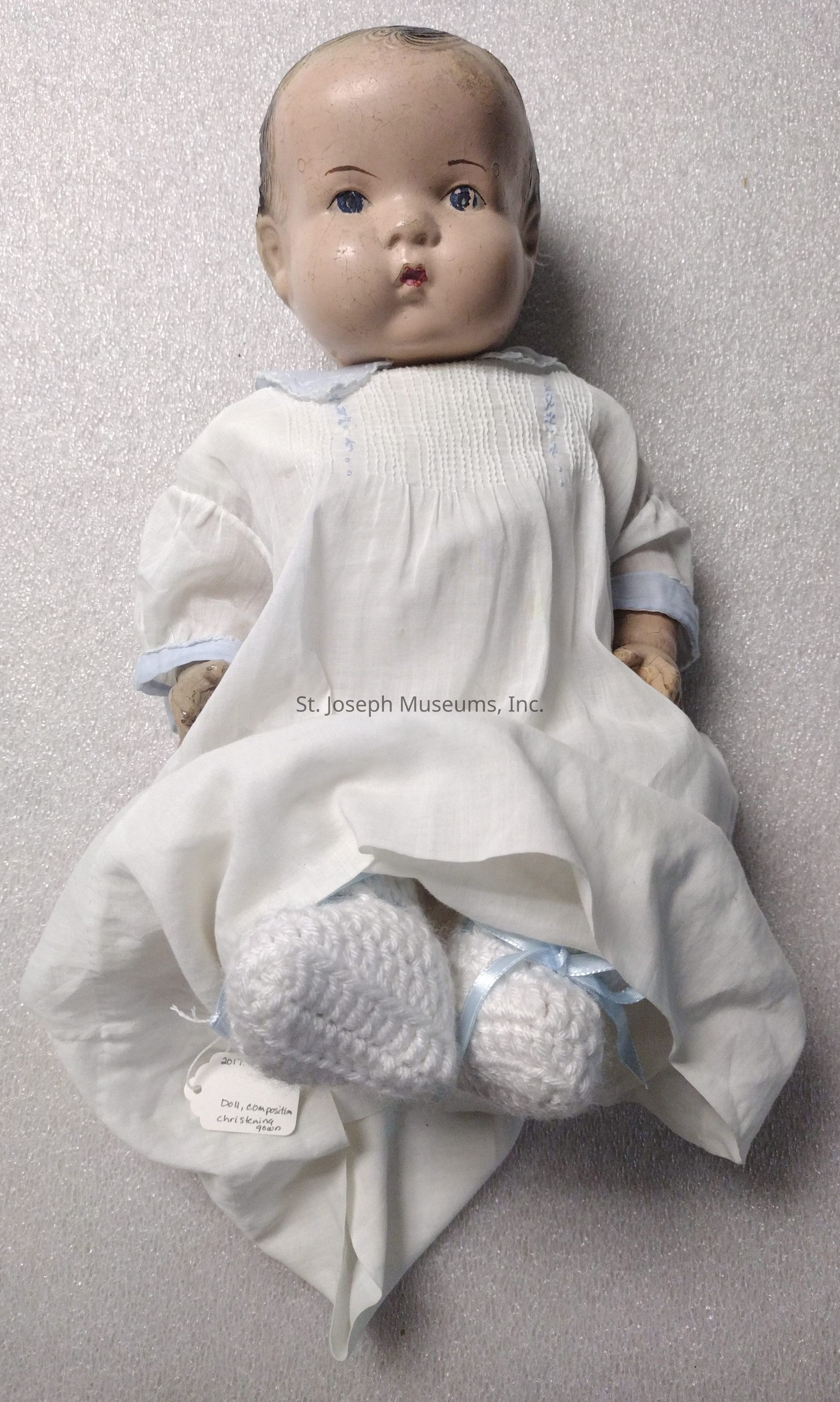 The image depicts a vintage composition doll dressed in a classic white christening gown, adorned with delicate embroidery details on the bodice, matched with soft, crocheted white booties tied with light blue ribbons, indicating a traditional and timeless aesthetic. The doll's face, with its subtly worn paint and a hint of cracking, reflects its age and adds a nostalgic charm, reminiscent of early to mid-20th-century craftsmanship. Placed against a textured, neutral gray background, the doll stands out, giving a sense of fragility and history, while also evoking memories of bygone childhoods. A small tag is visible, suggesting its classification as a cherished collectible item, further emphasizing its antique nature and the sentimental value often associated with such heirloom pieces.