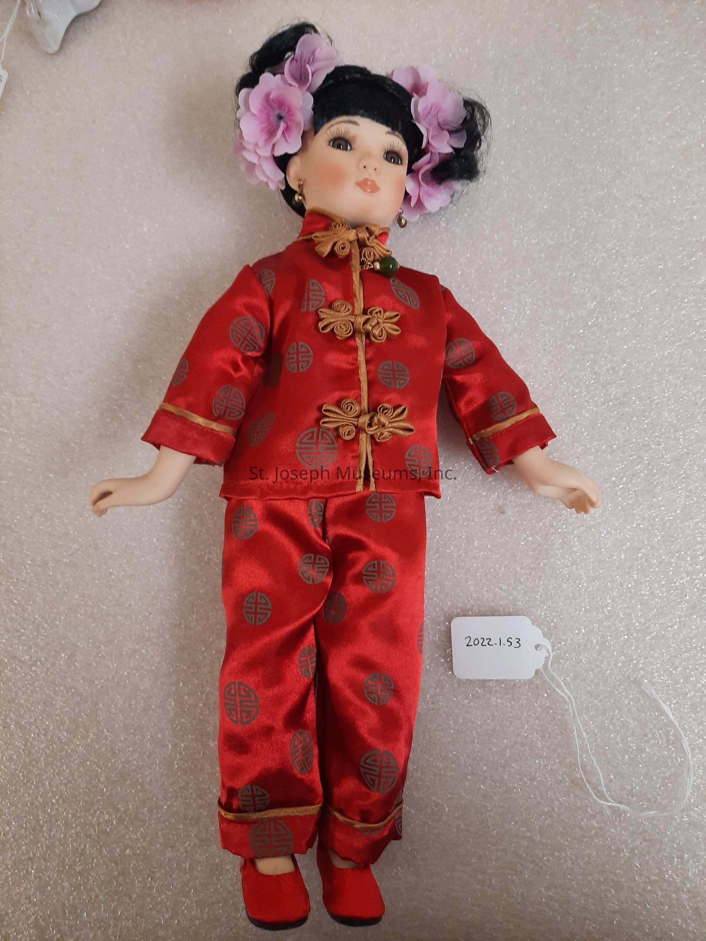 The image depicts a porcelain doll dressed in a traditional red satin outfit adorned with golden knots and circular patterns, reflecting a cultural or festive style. The doll has a serene expression, with its hair styled in a sleek fashion, adorned with large, pink floral accessories. The clothing consists of a long-sleeved top and matching pants, showcasing a consistent design motif. The doll also features red shoes that complement the ensemble. Additionally, it wears earrings, adding an elegant touch to its appearance. A small tag labeled "2022.1.53" is attached, indicating cataloging or collection details. The background is a simple textured surface, focusing attention on the doll.