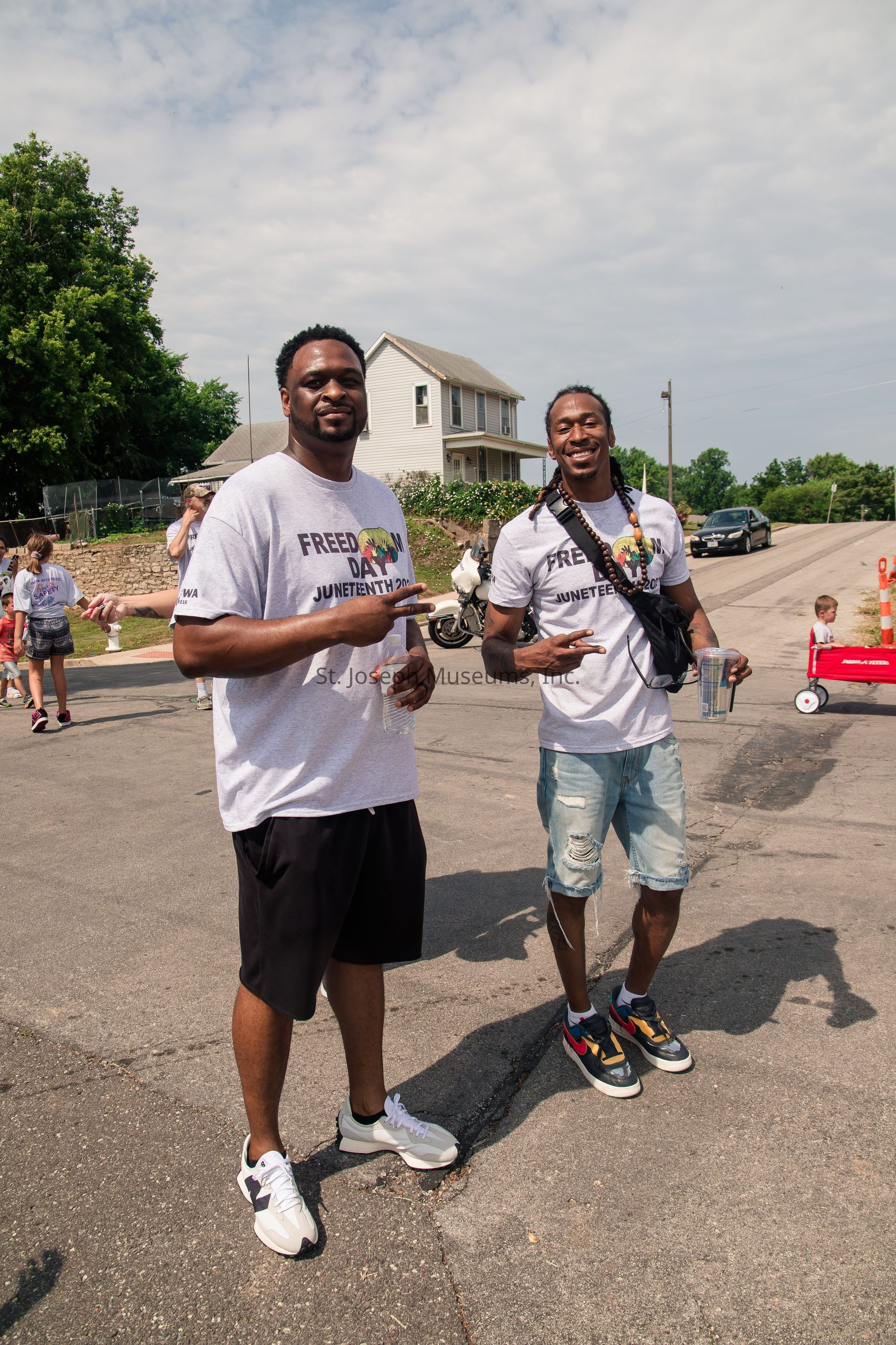 The image depicts two men standing on a street during a community event, both wearing matching "Freedom Day Juneteenth 2021" t-shirts, with one in black shorts and the other in ripped denim shorts; they are smiling, holding drinks, and making peace signs at the camera, while in the background, several people, including a child in a red Radio Flyer wagon, are visible, along with a house, greenery, and a car parked on the side, all under a partly cloudy sky that enhances the festive, relaxed atmosphere of the scene.