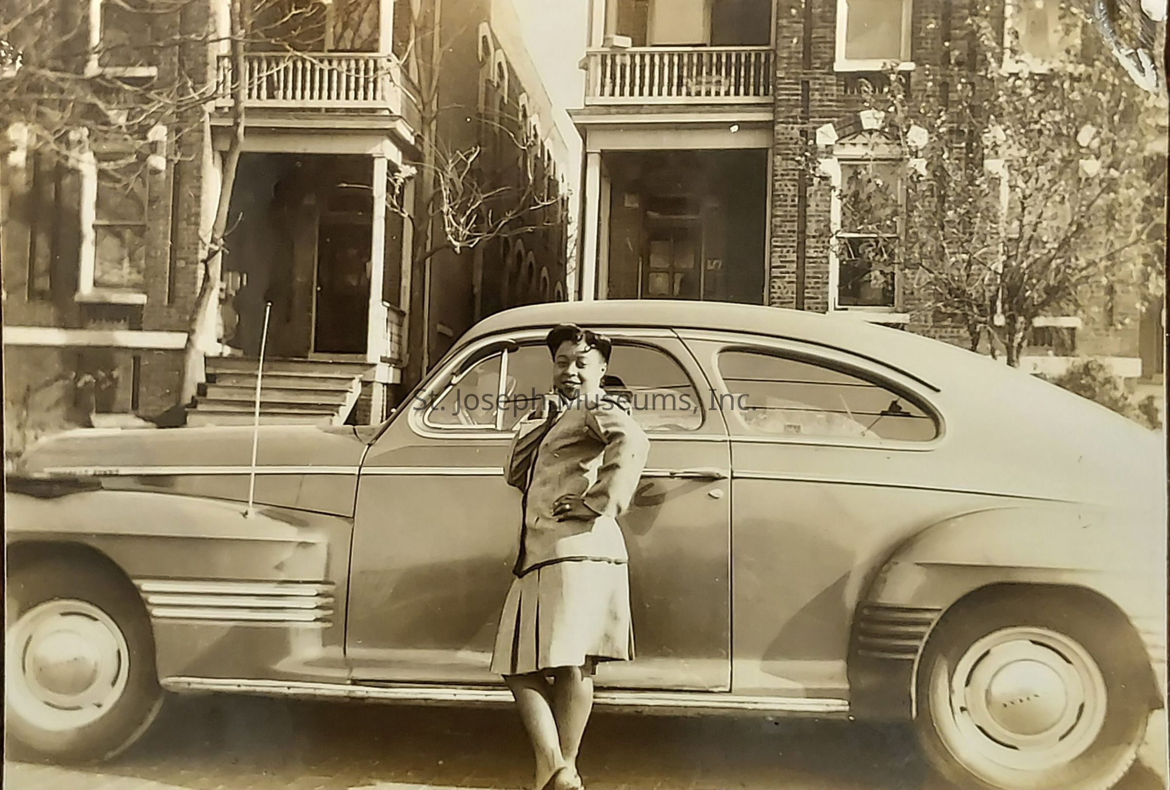 The image depicts a vintage scene in which a woman stands confidently beside a classic car, possibly from the mid-20th century, parked in front of a row of brick houses. She is dressed in attire typical of the era, wearing a skirt suit with a jacket and pleated skirt. Her pose, with one hand on her hip and a smile, suggests a sense of pride, perhaps in the car or the moment. The car, with its rounded lines and chrome details, reflects the design trends of its time. The background consists of urban residences, characterized by brick walls, wooden steps, and visible metal railings. Leafless trees suggest the photo was taken in a colder season. Overall, the black and white photograph captures a nostalgic moment, highlighting the style and atmosphere of the period, while evoking a sense of timelessness and personal history.