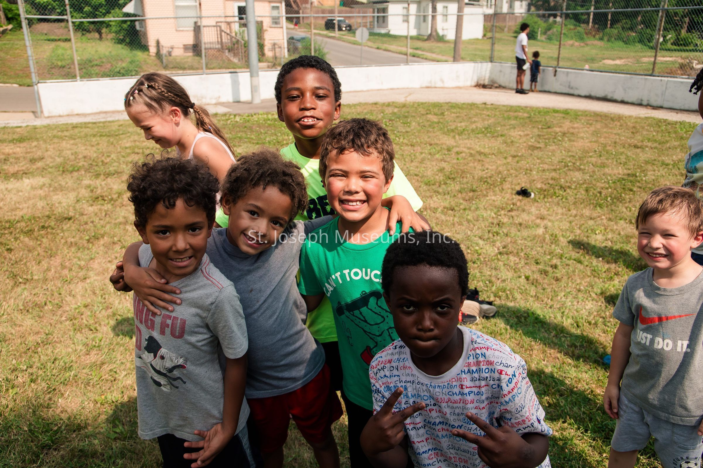 In this vibrant image, a group of young children are gathered outdoors on a sunny day, standing closely together on a grassy field. The kids, all smiling joyfully, are dressed casually and seem to be enjoying each other's company. One child in the front is playfully making a peace sign with his hands, while others are huddled in a friendly embrace. In the background, a couple of other children can be seen near a chain-link fence that surrounds the area, while some residential houses are visible in the distance. The scene exudes a sense of camaraderie and carefree fun.
