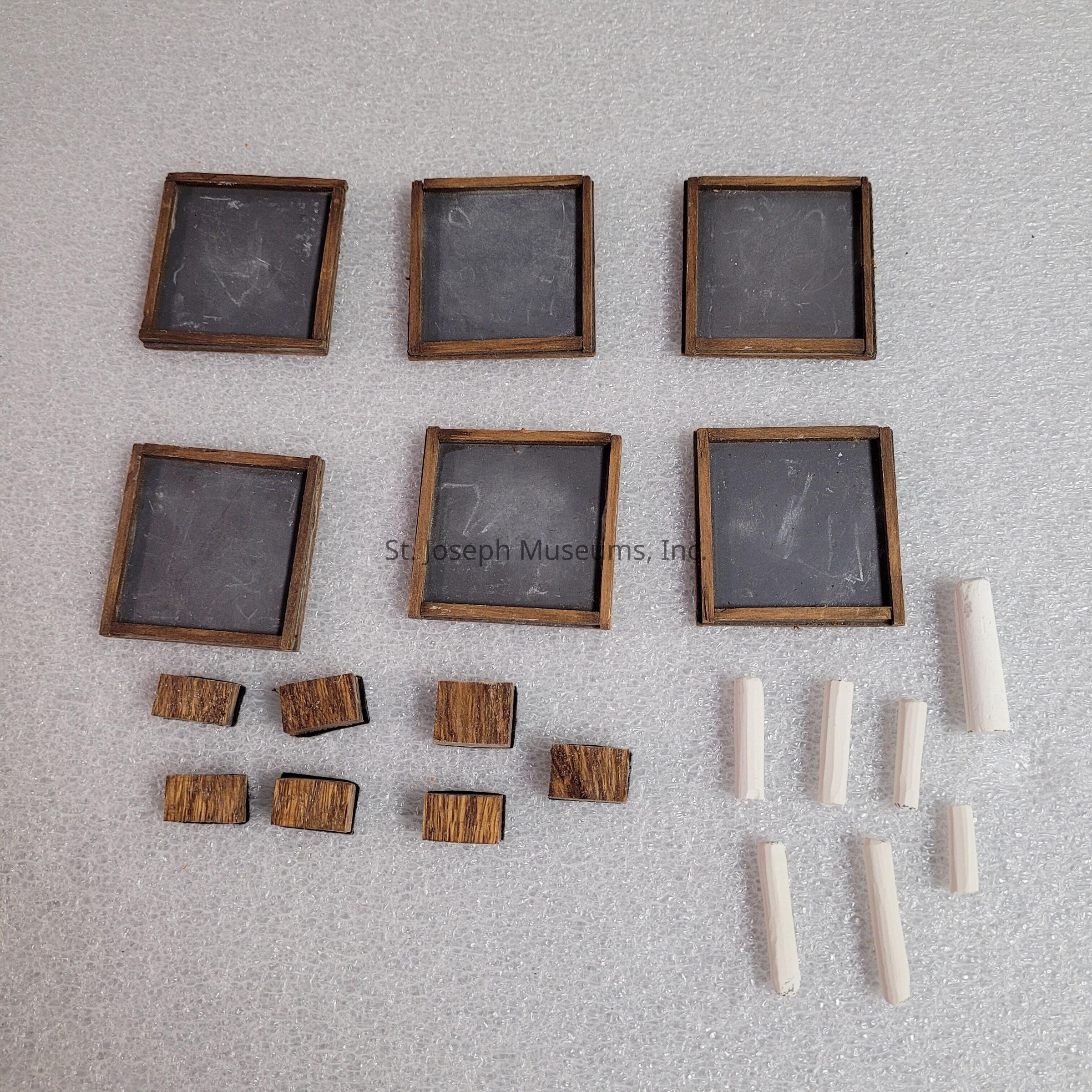 The image displays a collection of small square wooden slate boards, seemingly arranged in two rows of three, each bordered by a rustic wooden frame. Below these slates, a series of eight small wooden blocks, square in shape, is arranged in two rows of four, appearing to be made from the same type of wood as the frames. Adjacent to the small wooden blocks, on the right side, a total of eight slender, cylindrical pieces of white chalk are neatly positioned, four on top and four on the bottom, contrasting sharply against the textured, pale gray background on which all the items rest. This entire setup gives an impression of vintage educational tools or perhaps components of a small craft project, providing a sense of nostalgia and simplicity.