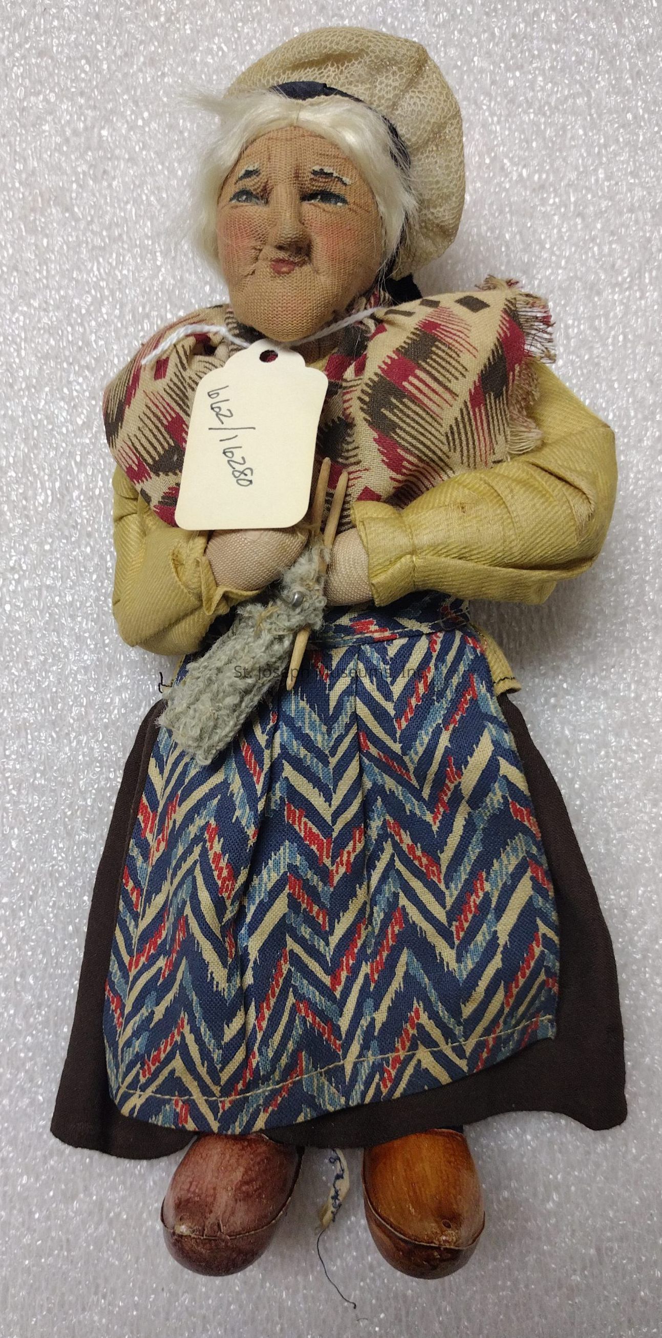 The image depicts a handmade doll representing an elderly woman, characterized by a detailed and textured face with a gentle expression. She wears a traditional outfit, including a yellow blouse and a skirt adorned with a vibrant, multicolored chevron pattern, complementing the brown fabric. Her shoulders are draped with a patterned shawl, adding to her rustic appearance, while her head is covered with a beige bonnet. In her hands, she holds knitting needles with yarn, suggesting an engaged and industrious lifestyle. Her footwear consists of wooden clogs, enhancing the authenticity of her attire. A paper tag attached to her shawl bears a handwritten note or code, adding a personalized element to the figurine. The doll seems to embody cultural or historical significance, possibly representing a specific region or traditional craftsmanship. Positioned against a textured, light-colored backdrop, the doll’s features and clothing are accentuated, providing a vivid and lifelike portrayal, capturing the viewer’s attention with its intricate craftsmanship and nostalgic charm.