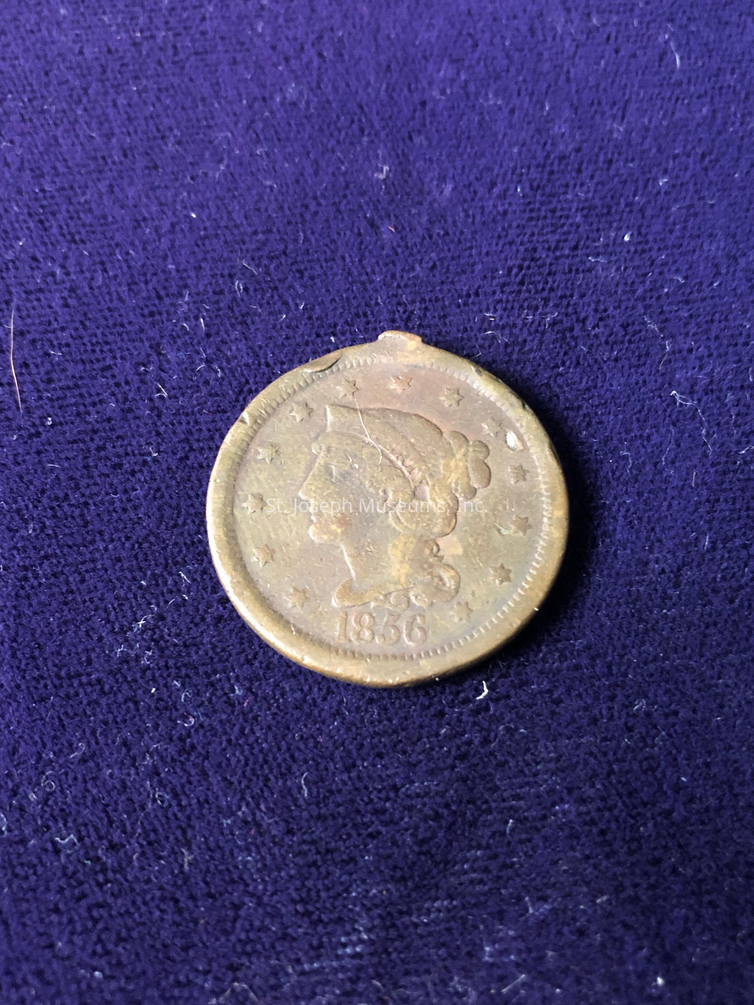 The coin, dated 1856, appears to be from the United States and exhibits a woman’s profile surrounded by stars along the edge, indicative of the Liberty Head design used during that period. The surface of the coin displays a patina, characteristic of its historical age, with subtle signs of wear that hint at its long existence. The textile underneath adds texture to the composition, with visible fibers contrasting the smooth metallic surface of the coin, enriching the visual appeal and context of the scene.