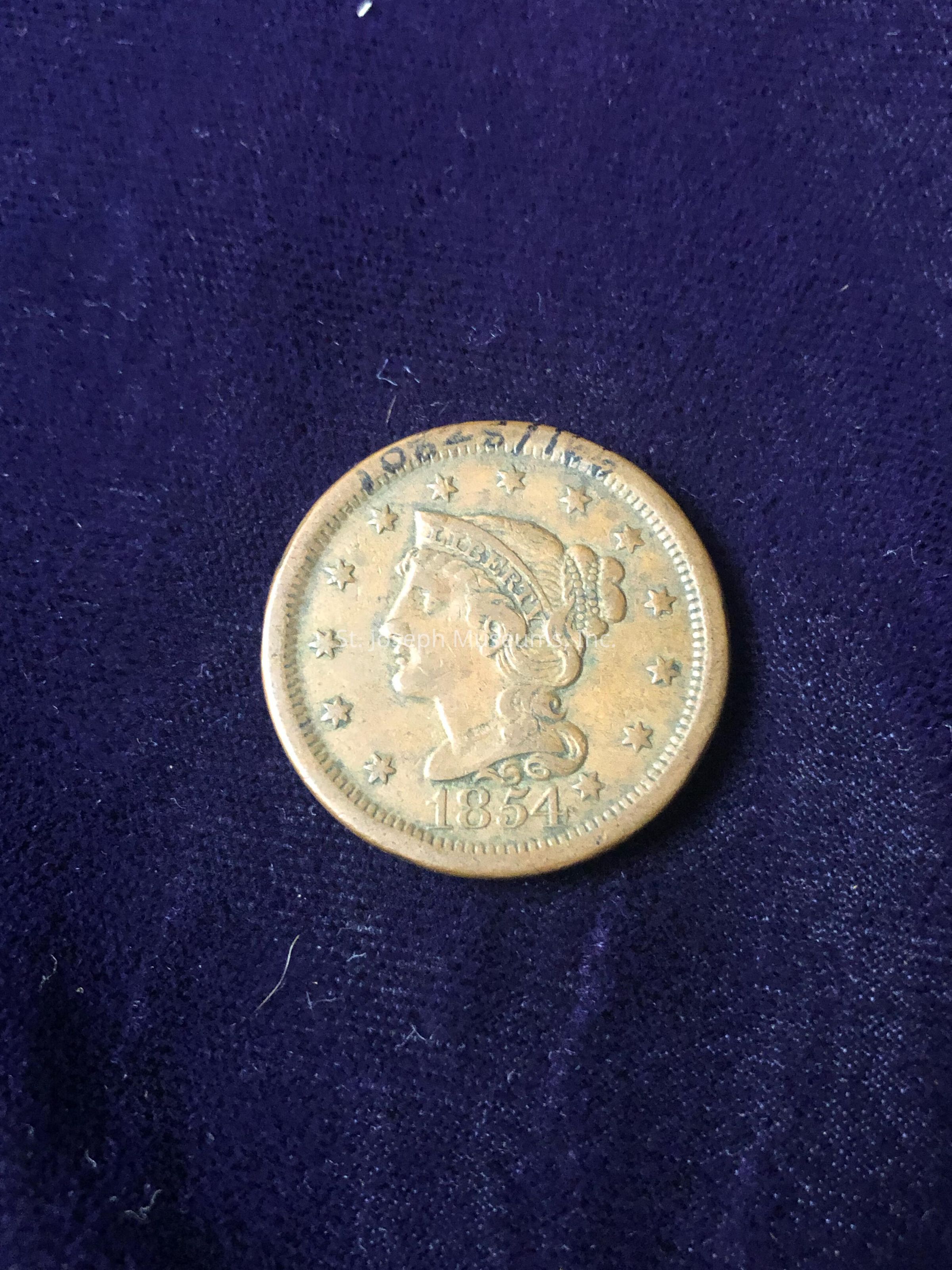 The image showcases an antique coin from the year 1854, featuring a profile of Lady Liberty adorned with a headband that reads “LIBERTY,” and surrounded by 13 stars signifying the original American colonies; the coin has an ornate design with Liberty’s hair tied back as a small bun pinned with a decorative piece, conveying a sense of historical elegance, and the visible date along the lower edge further emphasizes its vintage origin; this numismatic artifact is laid on a textured dark blue or purple cloth that contrasts with the coin’s aged golden hue, highlighting its intricate engraving and well-preserved state, suggesting the coin may be of significant collectible value, as it carries not only historical worth but also exemplifies the artistry of 19th-century coinage, and altogether, the image captures a moment of American monetary history, inviting contemplation about the coin’s journey through time while resonating with the present as a tangible connection to the past, thereby evoking a blend of curiosity and appreciation for history enthusiasts