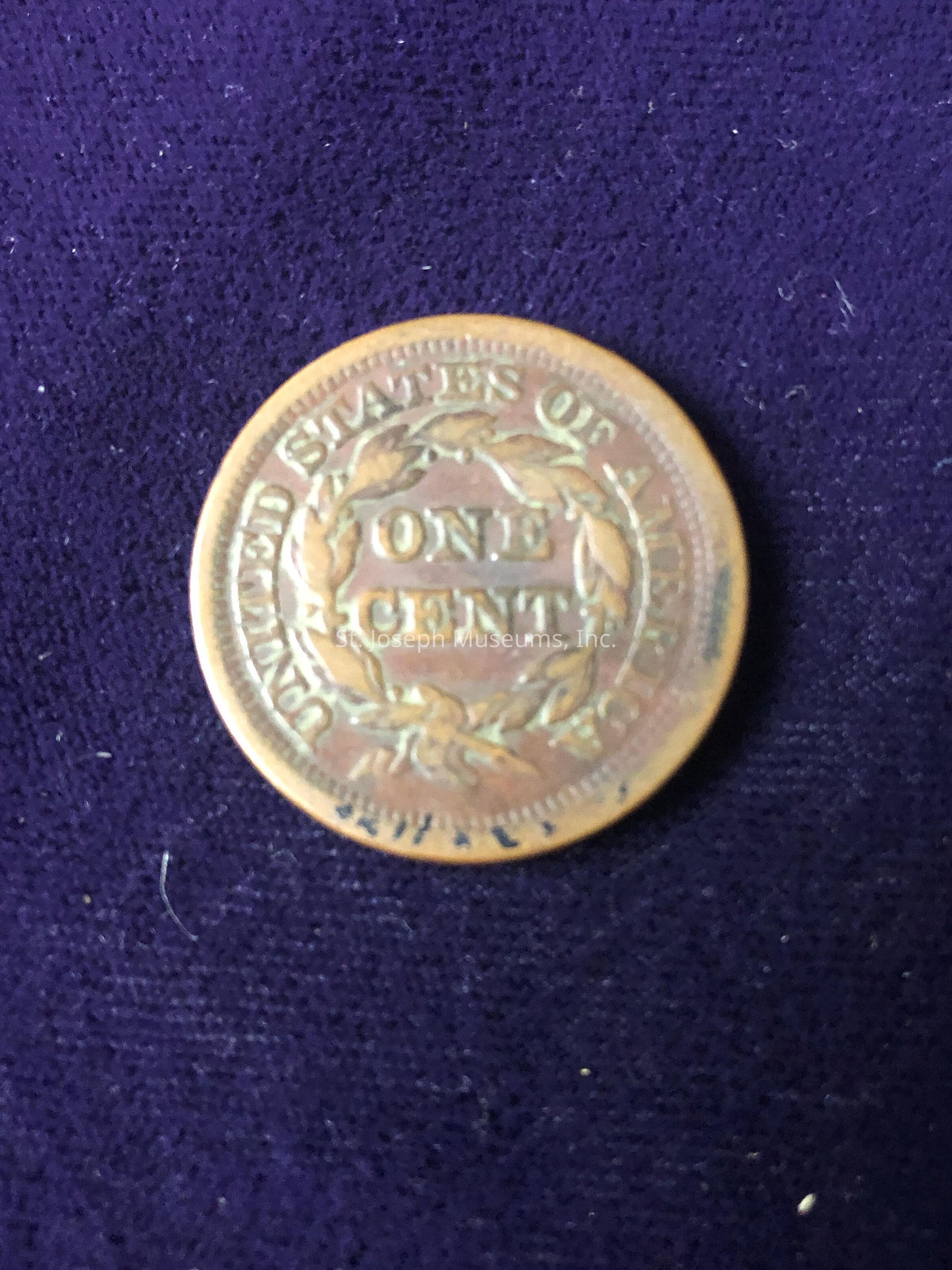 The coin features the inscription "ONE CENT" encircled by a laurel wreath with the words "UNITED STATES OF AMERICA" along the outer edge. The coin exhibits a warm, bronzy patina, indicative of its age and historical value. Details of the coin’s engravings are accentuated by its worn, slightly tarnished surface, suggesting a long history, possibly belonging to a numismatic collection. The coin’s distinct textures and colors are highlighted against the contrasting background, emphasizing its vintage appeal.