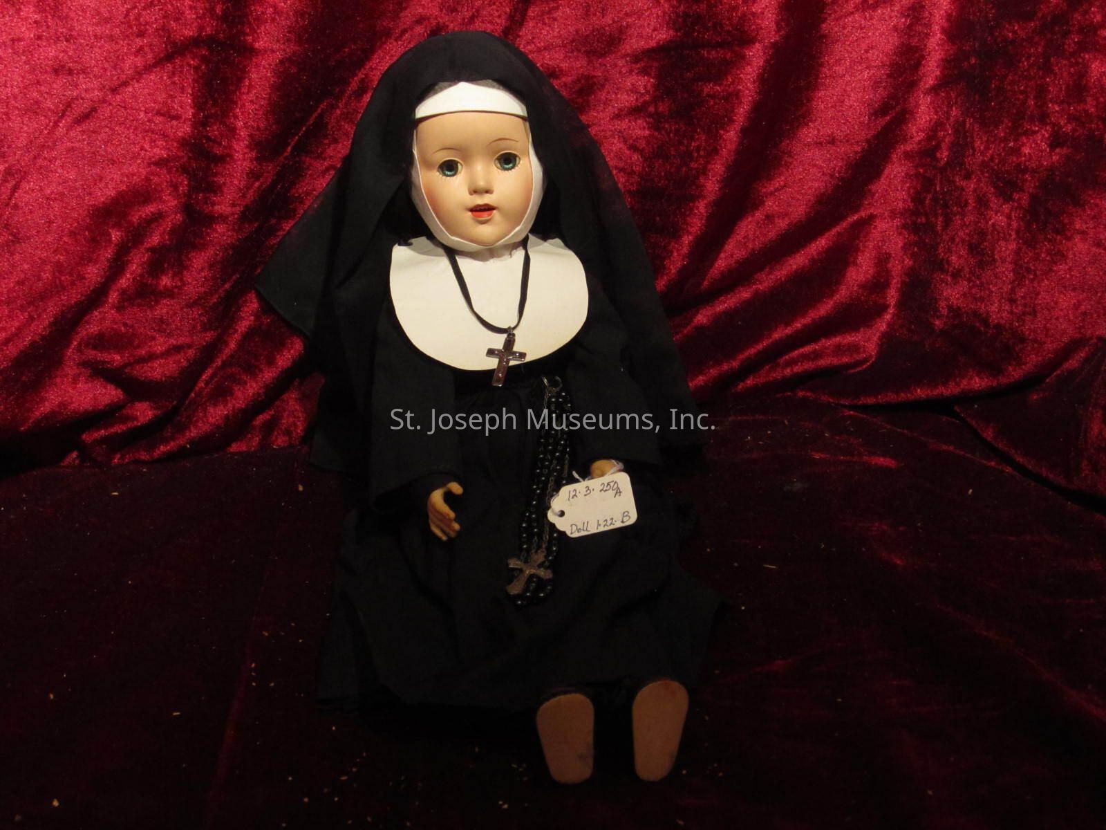 12.3.250 A: The image depicts a doll dressed as a nun, adorned in a traditional black habit with a white wimple and veil. The doll is seated against a rich, red velvet backdrop, which adds a dramatic contrast to its attire. Around its neck hangs a rosary with a crucifix, and a small tag is attached to its garment, inscribed with what appears to be a date or reference number. The expression on the doll's face is serene, reflective of its religious theme, and the overall presentation suggests an antique or collectible item, carefully displayed to emphasize its features and attire.
