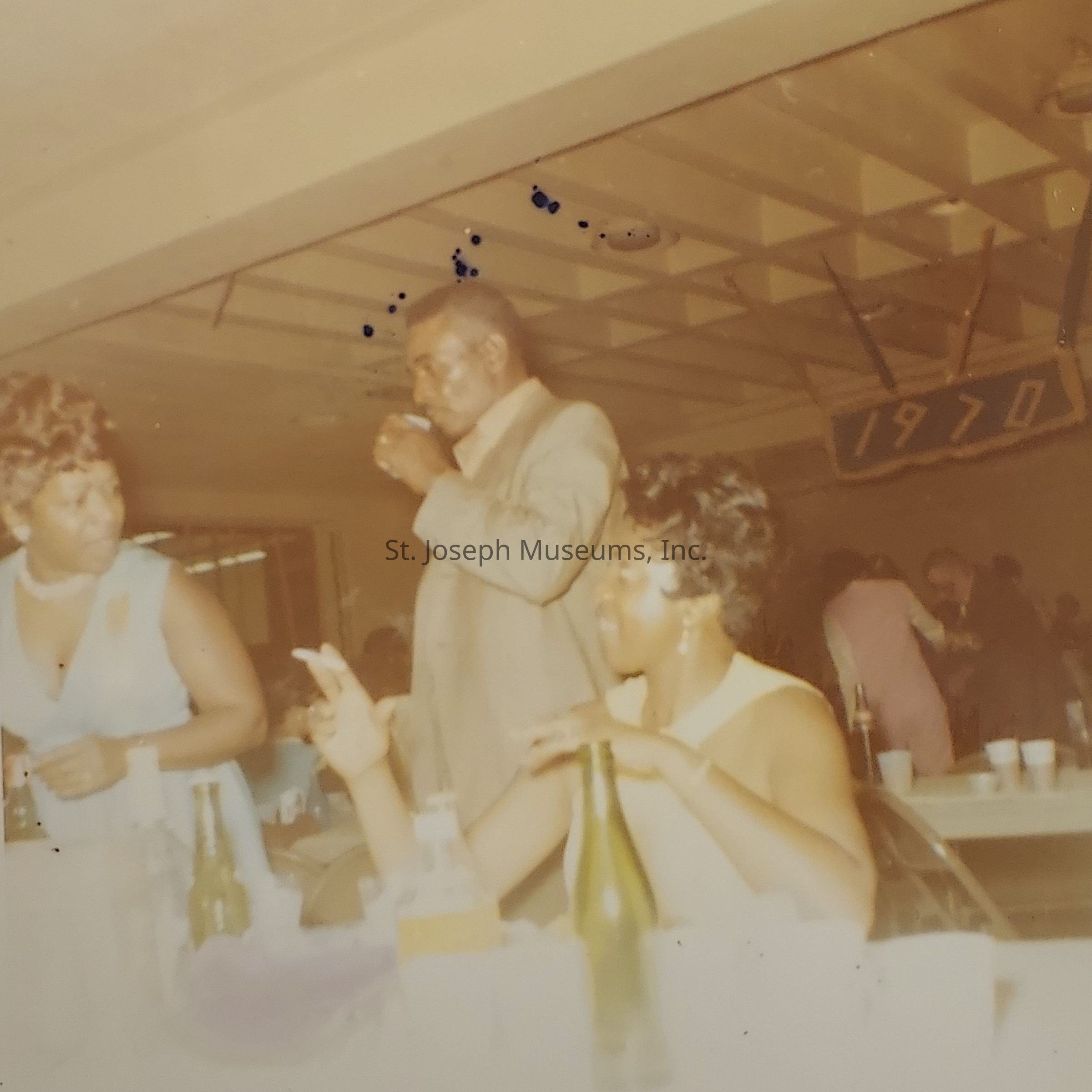 The image captures a lively indoor gathering from the year 1970, as indicated by a sign on the wall. In the foreground, two women and one man are engaged in conversation at a table covered with bottles and cups, suggesting a social or celebratory event. The woman on the left, adorned in a light blue dress, appears to be speaking animatedly to the seated woman in a yellowish dress, who is gesturing with her hands. The man standing nearby is wearing a suit and drinking from a cup. The background shows more attendees enjoying the occasion, highlighting a warm, social atmosphere.
