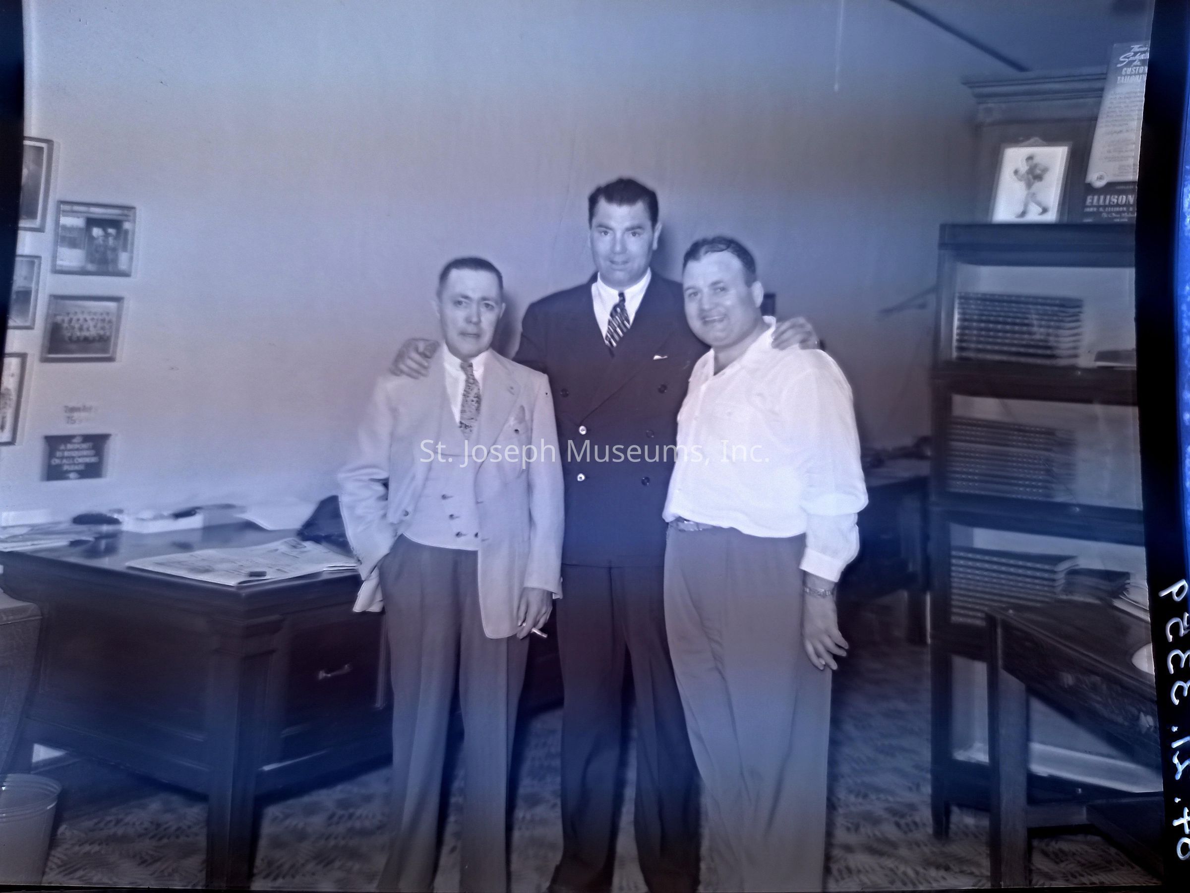 Black-and-white image of three men standing together inside an office at 118 North 8th St., Joseph, Missouri. The men have been identified as Jack Dempsey with Lawrence Weldon and Dr. Fordyce. The man in the middle, Dempsey, is dressed in a dark suit, while the two on either side wear lighter-colored suits, one with a noticeable vest and tie. They stand by a large wooden desk with neatly arranged papers and shelves filled with documents.