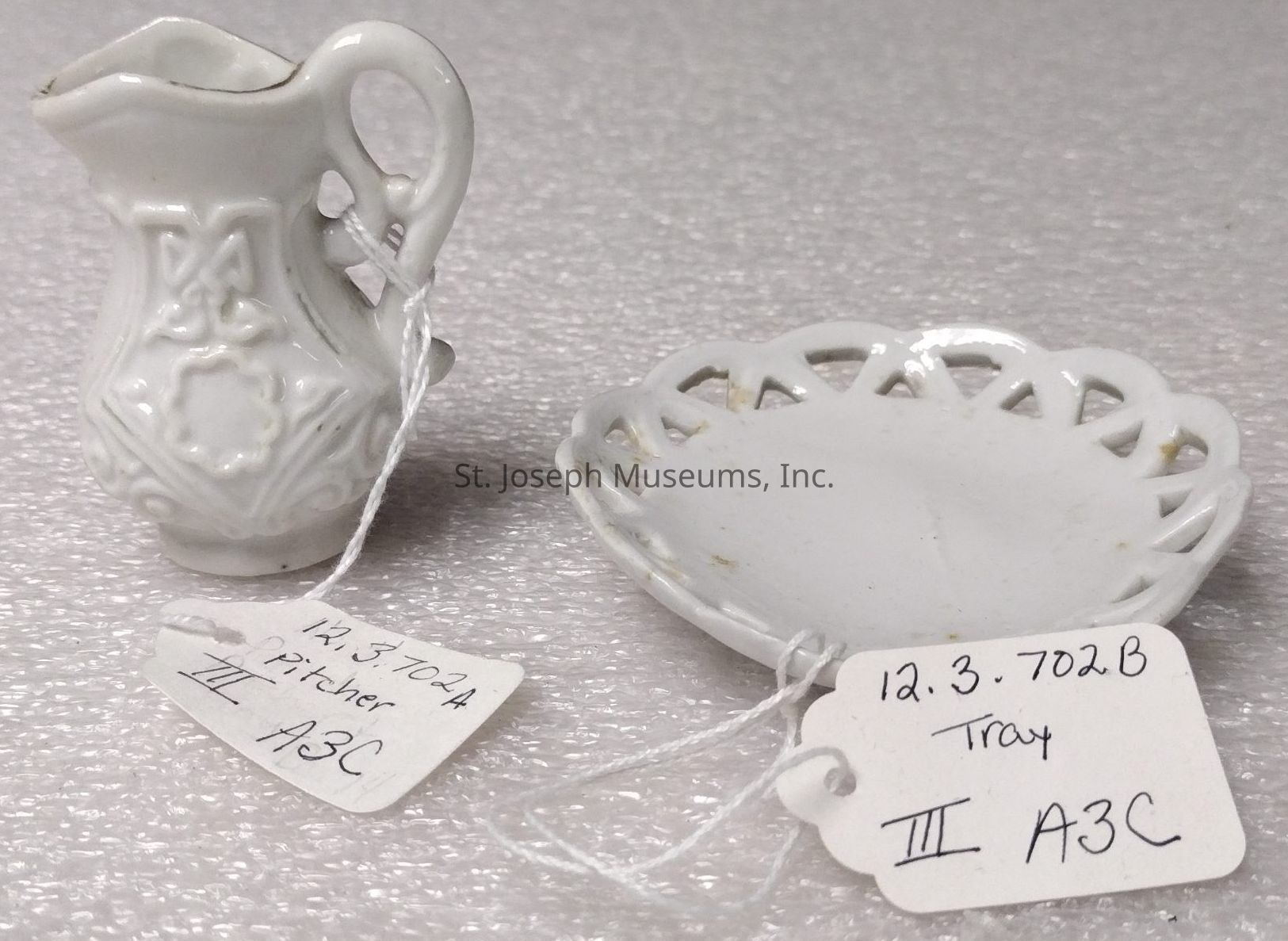 The image displays two miniature porcelain items on a textured surface: a delicately crafted white pitcher with ornate embossed designs and a handle, alongside a matching round tray with a slightly elevated, lattice-like rim. Both items have paper tags attached, secured with strings; the tag on the pitcher reads "12.3.702A Pitcher III A3C," while the tray's tag shows "12.3.702B Tray III A3C." These details suggest they are part of a categorized collection, possibly for display or archival purposes, reflecting a vintage or collectible aesthetic.