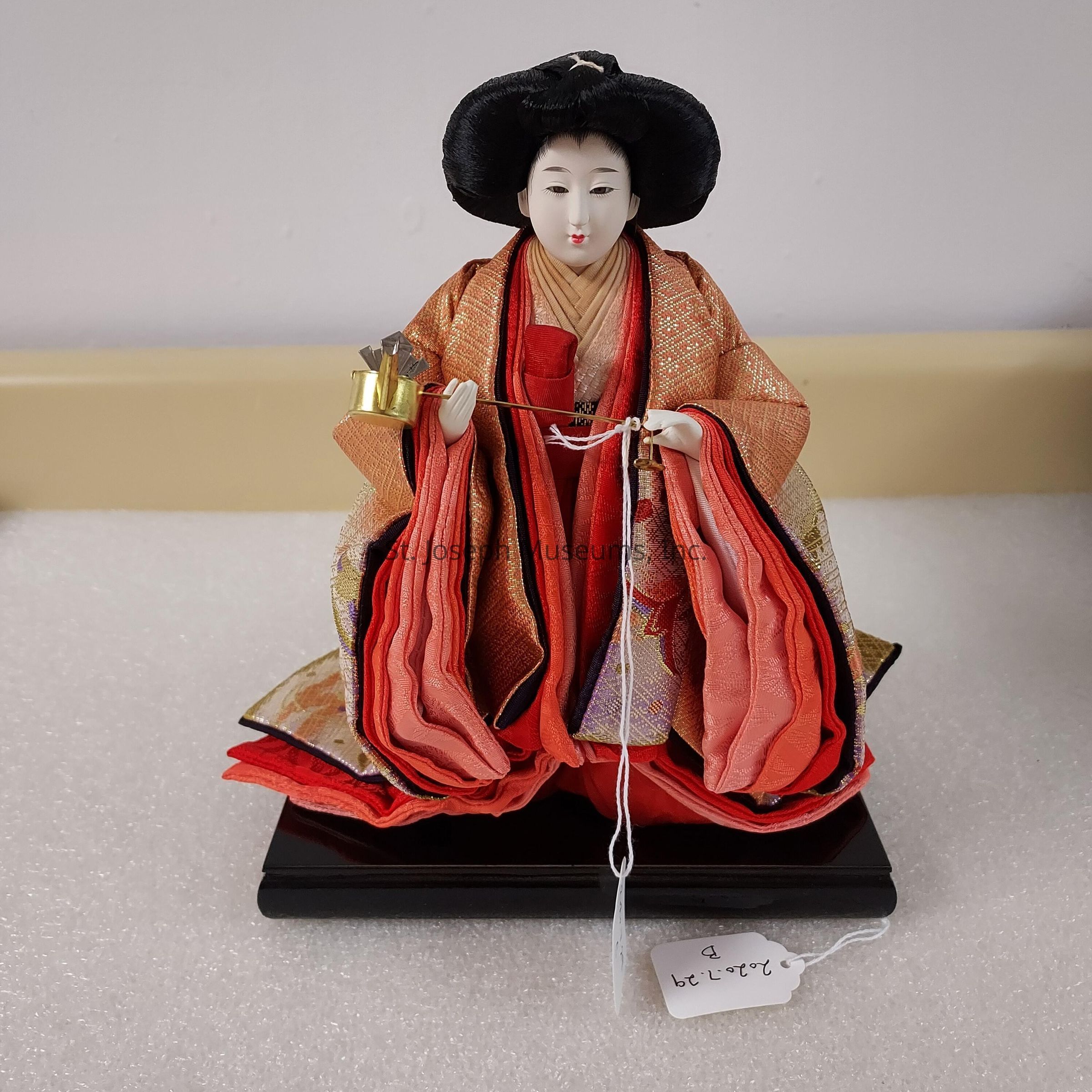 The image features an exquisite Japanese doll dressed in an elaborate, traditional kimono. The doll is adorned with layers of rich fabric in shades of orange and red, meticulously crafted with intricate patterns and fine textures. Her black hair is styled elegantly, and she holds a delicate object in her hands, possibly a small musical instrument or ceremonial item. The doll's expression is serene, with finely painted features that convey grace and refinement. It is positioned on a black pedestal, and there is a small tag attached to it with some handwritten numbers or letters. The background is simple, emphasizing the doll's intricate craftsmanship and cultural significance.