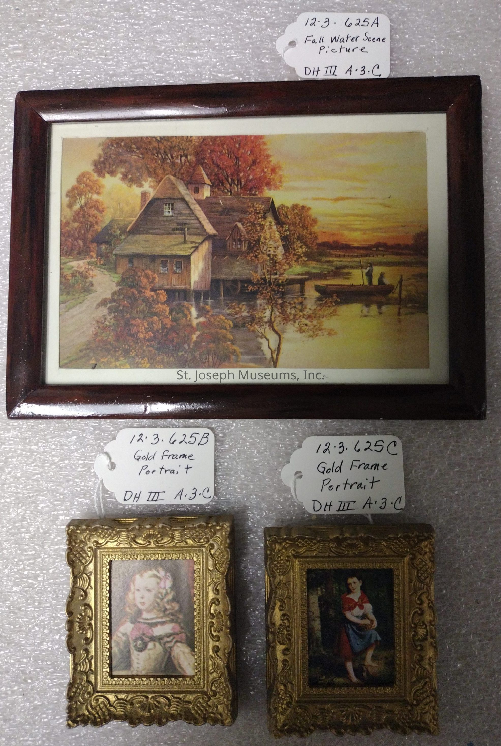 The image displays a collection of three framed artworks against a textured background. The top piece is a framed picture of a picturesque autumn water scene with a wooden house surrounded by vibrant fall foliage, labeled "12.3.625A Fall Water Scene Picture" in handwritten text on a tag. Below are two smaller gold-framed portraits, each featuring detailed ornate designs. The left portrait depicts a person in historical attire and is labeled "12.3.625B Gold Frame Portrait," while the right shows a figure in a seated pose, labeled "12.3.625C Gold Frame Portrait." Both tags share the annotation "DH III A.3.C." These diverse frames create a nostalgic and classic display.