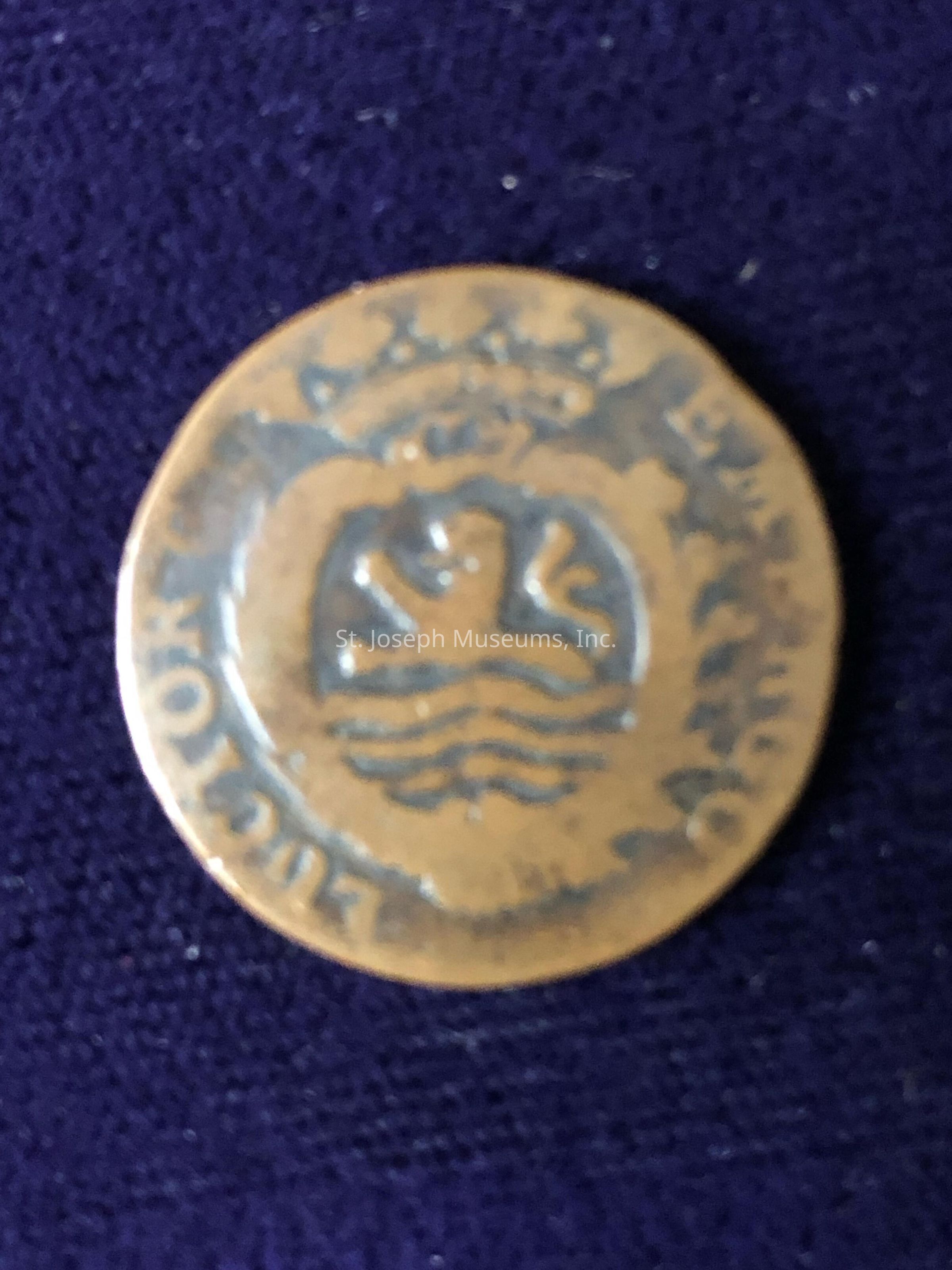 The object appears to be made of metal, featuring a worn, possibly tarnished surface, with a central design consisting of a heraldic emblem that includes a lion and wavy lines, enclosed within an ornate circular border. Above the central design is a depiction of a crown, indicating a possible royal or official insignia. Around the medallion, there is text inscribed in a circular manner, though some of it is blurred or worn, making it difficult to fully decipher. The overall appearance suggests an artifact with historical or cultural significance, possibly hailing from a certain organizational or geographical entity, and likely reflects the craftsmanship characteristic of a past era, providing a sense of aged authenticity and intriguing mystery.