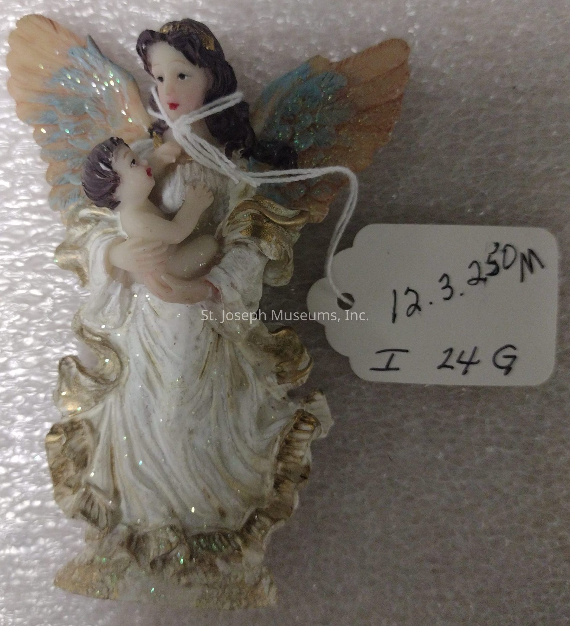 The image displays a decorative figurine of an angelic figure holding a child, both crafted with intricate detail. The angel features delicately painted features, wearing a flowing, glitter-adorned garment with gold accents and translucent, shimmering wings in shades of blue and peach. The child appears to be reaching towards the angel with a joyful expression. A string is tied around the figure, attaching a white tag with handwritten text reading “12.3.250M” and “I 24 G.” The figurine is set against a textured, white background that enhances its ethereal appearance.