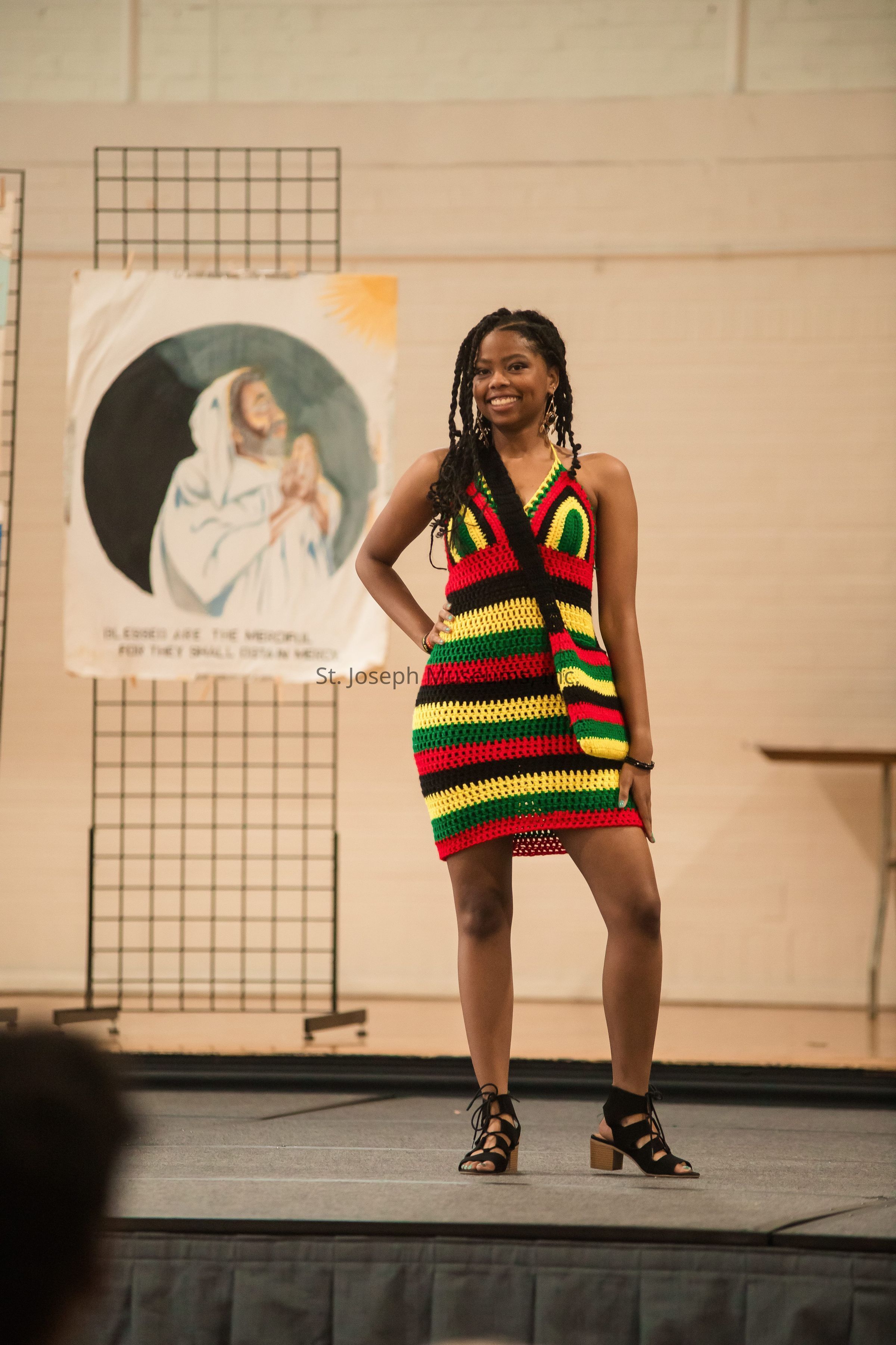 A woman stands confidently on a stage, wearing a vibrant crocheted dress with wide stripes of red, yellow, green, and black. Her hair is styled in long braids, and she is smiling warmly. She has one hand on her hip and is wearing stylish black sandals. Behind her, there is a backdrop featuring an artistic image of a figure, possibly religious in nature, accompanied by a message about mercy. The setting appears to be indoors at what might be a community event or fashion show, suggested by the stage and the casual, yet expressive, atmosphere.