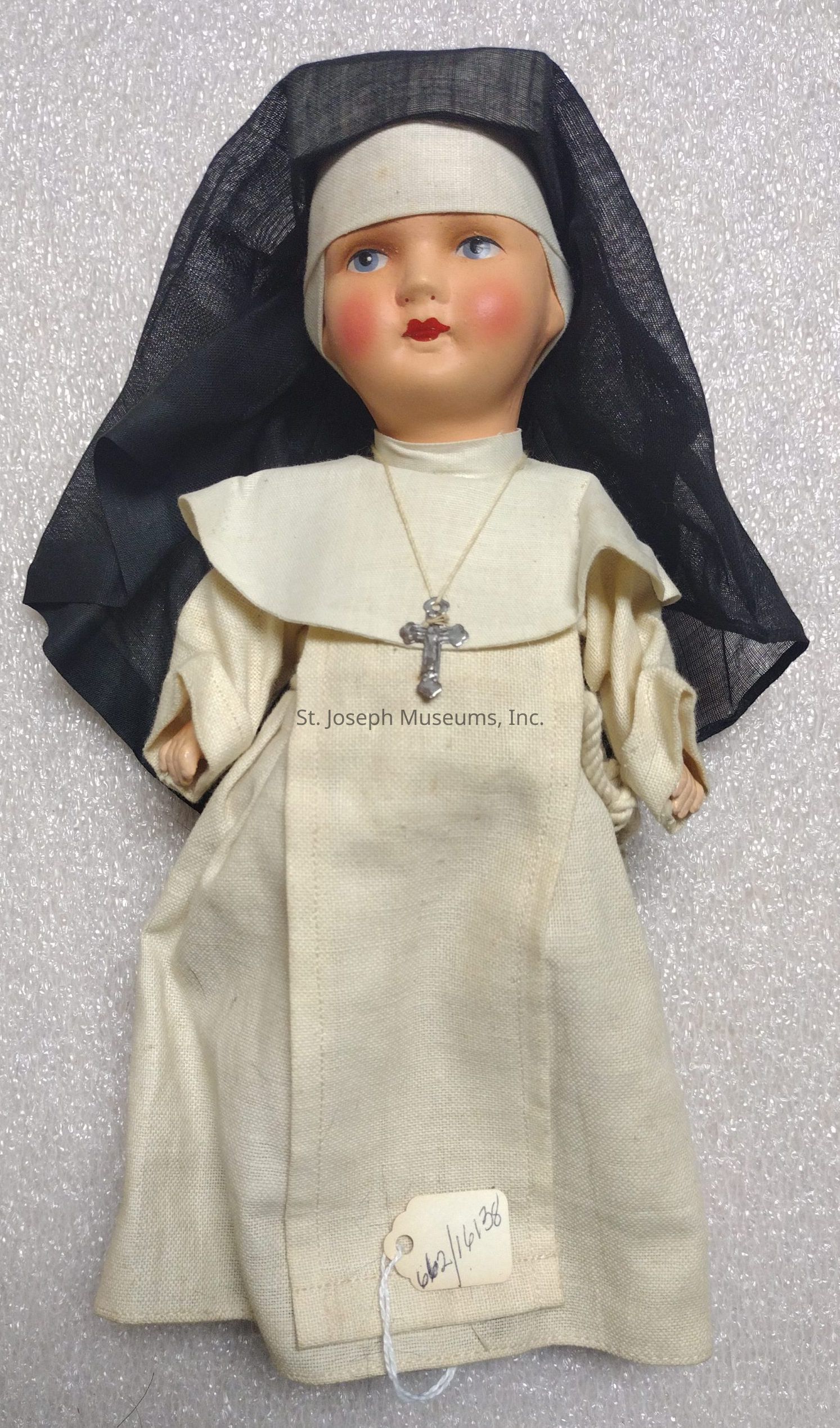 The image depicts a doll dressed in a traditional nun's habit. The doll is adorned in a cream-colored robe with a matching headpiece, along with a black veil covering the top and sides of the head. A silver cross necklace hangs around the doll’s neck, symbolizing religious devotion. The face has distinct features like a fair complexion, rosy cheeks, and striking blue eyes with red lips, giving it a serene and composed expression. The background is textured in a soft, neutral tone, and a small tag with handwritten numbers is attached to the bottom of the robe, suggesting it could be part of a collection or exhibit.