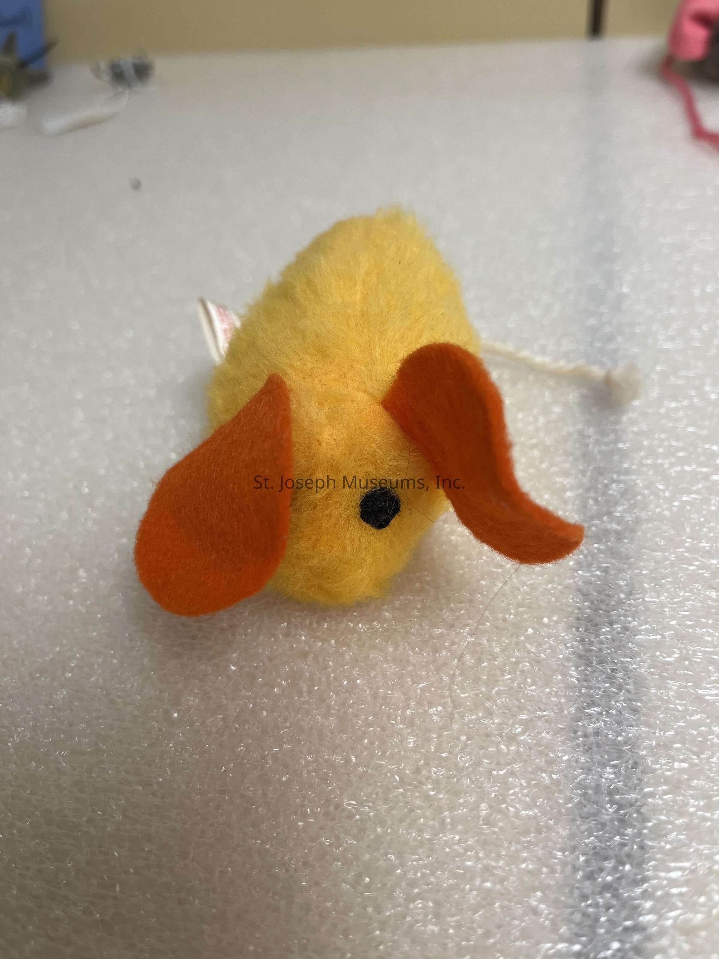 The image depicts a small, yellow, fluffy toy resembling a mouse, positioned on a textured white surface, with its two prominent orange ears standing out, and a small black nose adding to its cute and playful appearance, while a small tag is barely visible on the side, contributing to the impression that this is a handcrafted or unique piece possibly intended for decorative purposes or as a playful cat toy; additionally, the background shows some indistinct objects suggesting a workspace or crafting area, but the focus remains primarily on the charming little figure centered in the composition, capturing the viewer’s attention and sparking curiosity or imagination about its intended use or origin.