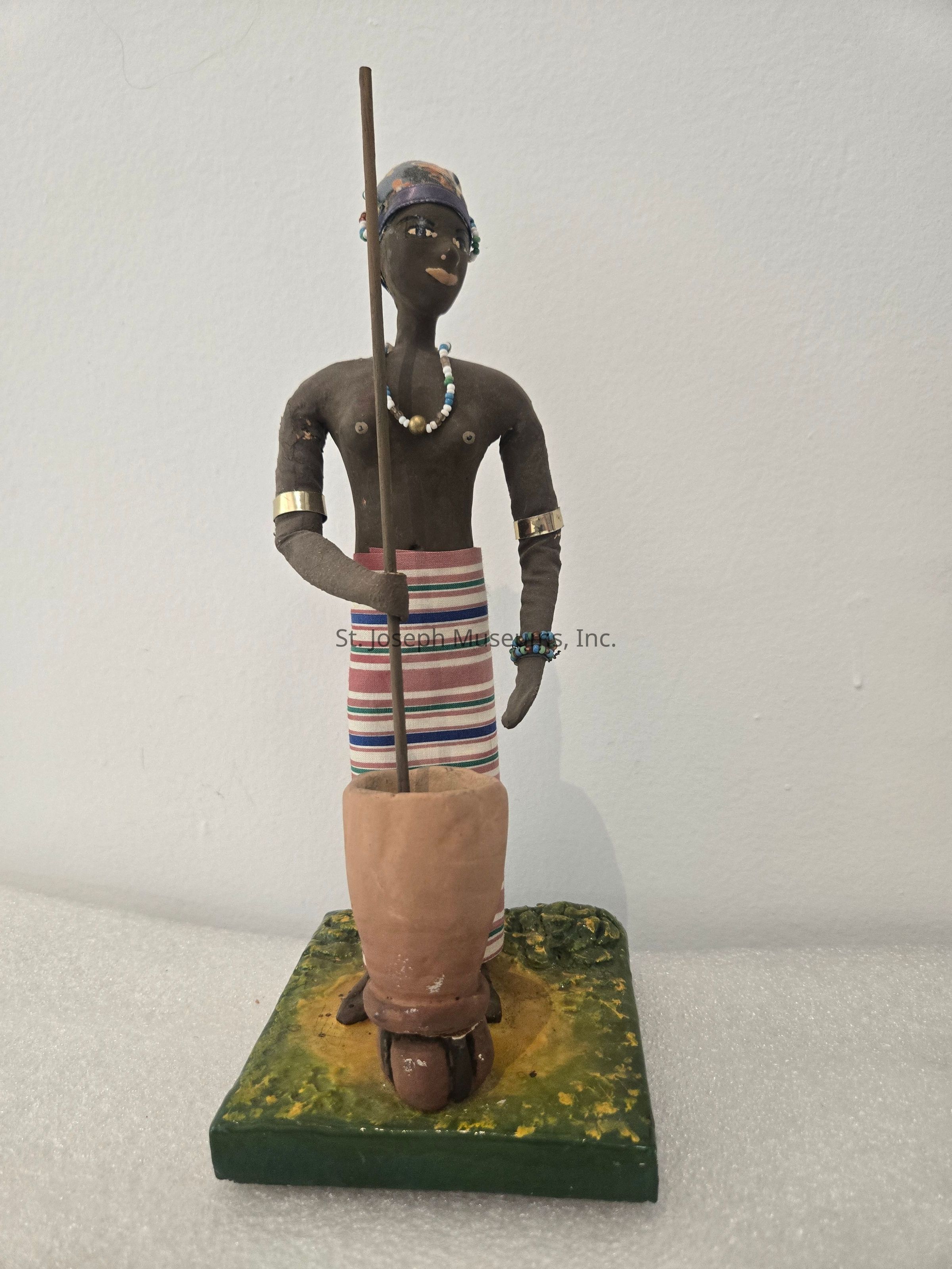 The image depicts a handcrafted figurine positioned on a flat, square base designed to resemble grass or moss. The figure is intricately detailed, wearing an elaborate headscarf and adorned with various beaded jewelry, like necklaces and bracelets, showcasing a vibrant array of colors. It is dressed in a cloth with horizontal stripes that wrap around the lower part of its body. The figurine holds a long stick, while standing beside a mortar, suggesting an activity like grinding or pounding, which could indicate cultural or traditional significance. Its arms are decorated with shining bangles, one on each wrist. The overall composition, with its skillful craftsmanship, and thoughtful use of color and form, offers a glimpse into cultural artistic expressions, highlighting attention to detail and cultural symbolism. The background is plain and white, focusing attention squarely on the figure, thus enhancing its presence and the storytelling aspect embedded within its posture and attire.