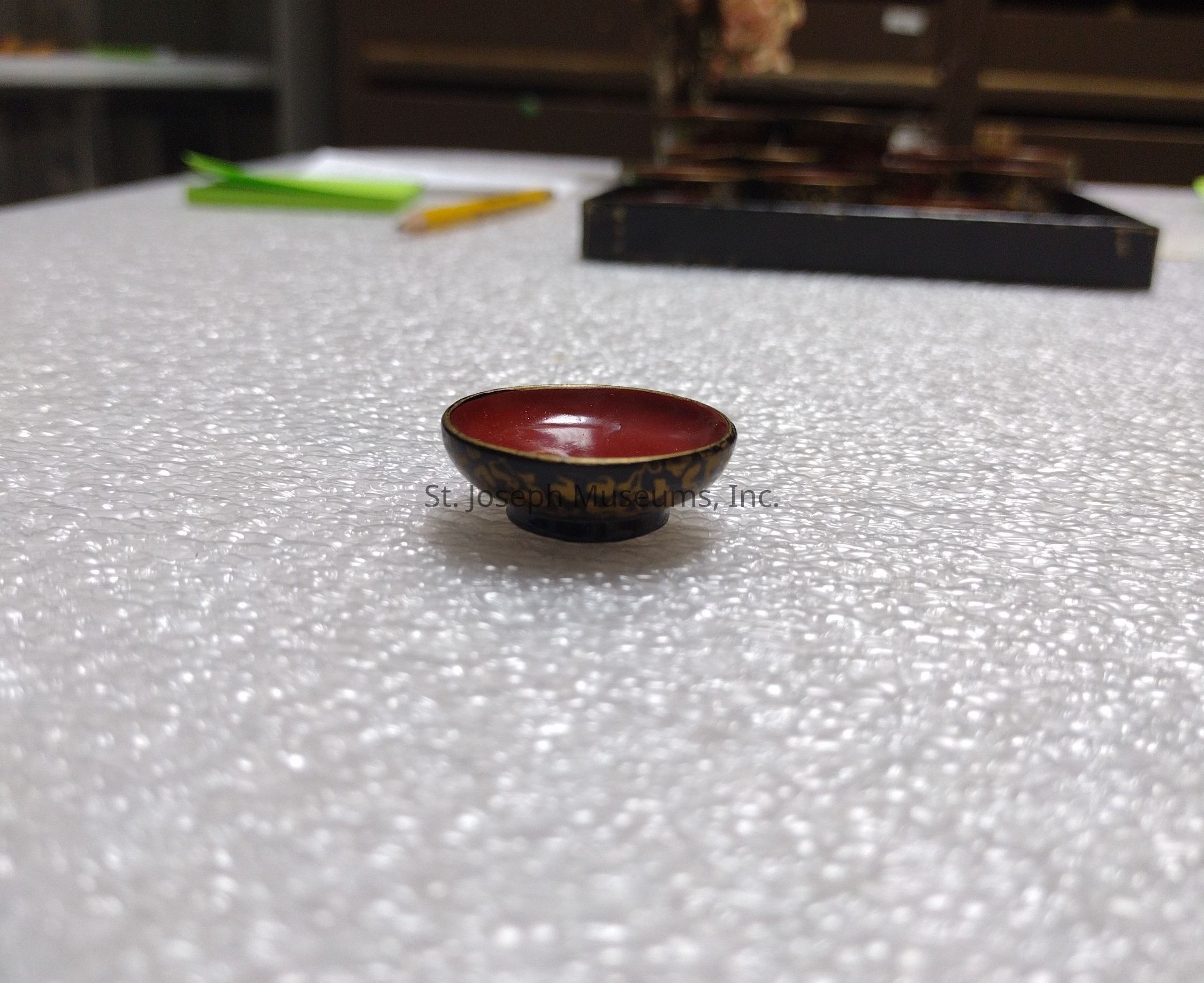 In the image, a small, ornately decorated bowl with a red interior and a patterned dark exterior sits on a textured, white surface reminiscent of bubble wrap. The background contains blurred elements such as a green piece of paper, a pencil, and what might be other similar bowls or objects. The focus remains on the bowl, emphasizing its intricate design and craftsmanship. The shallow depth of field highlights the bowl, leaving the background slightly out of focus, which draws the viewer's attention to the central object and its artistic detailing.