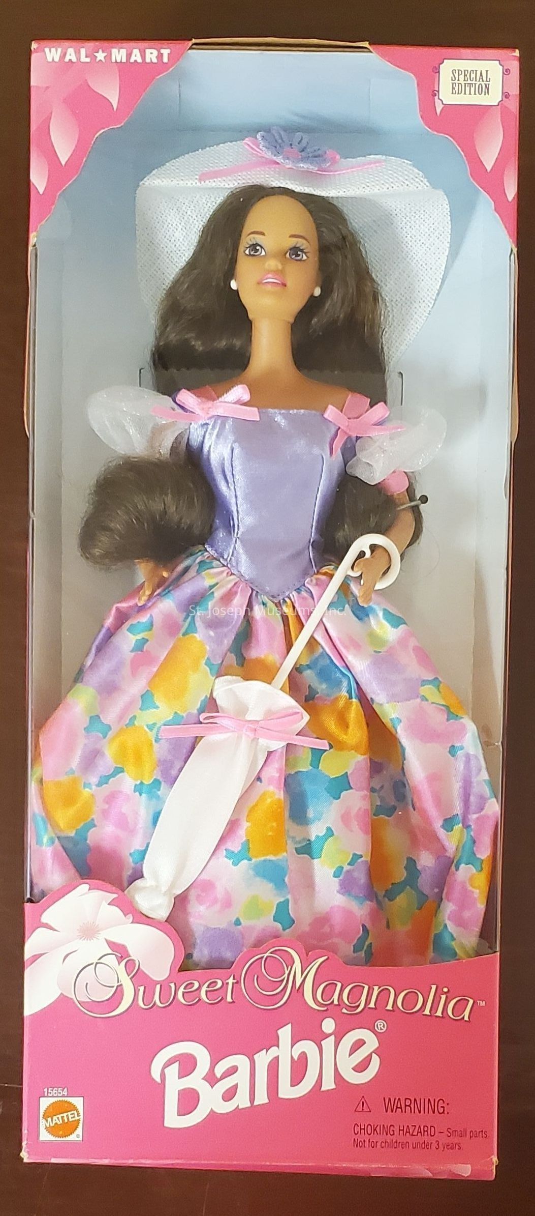 The image shows a "Sweet Magnolia" Barbie doll in its original packaging. The box is mainly pink with decorative floral motifs and the Walmart logo at the top. The special edition label is on the upper right corner. The doll is dressed in a colorful, floral-patterned dress with a pastel purple bodice adorned with pink ribbons on the shoulders. Her long, dark hair cascades down in soft waves, and she wears a wide-brimmed white hat embellished with a purple flower. She holds a delicate white parasol tied with a pink bow. The Mattel logo and product number "15654" are visible at the bottom, alongside a choking hazard warning for small parts not suitable for children under three years.