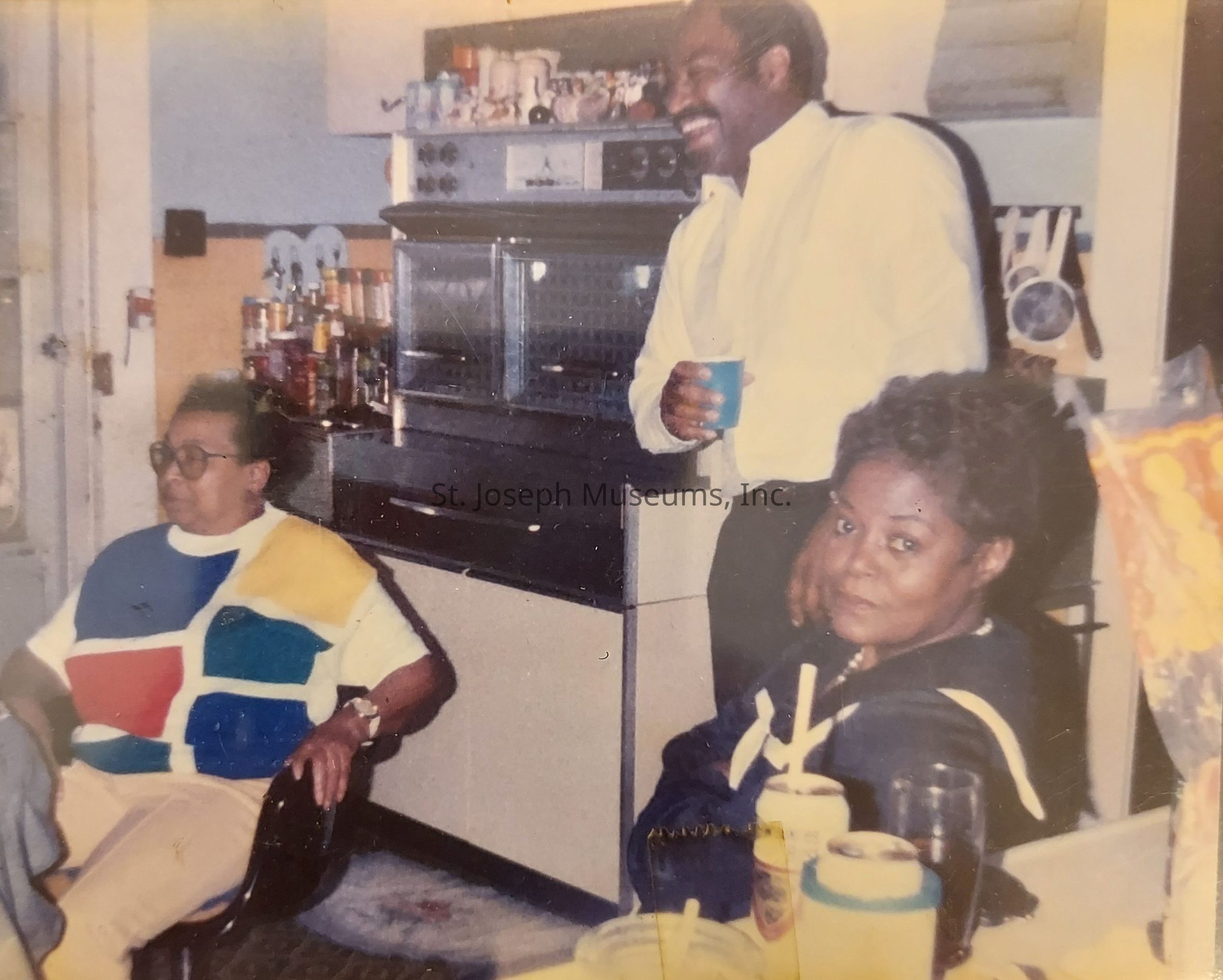 In the image, three people are gathered in a cozy kitchen setting. A person in the foreground is wearing a colorful shirt with a geometric pattern in blue, red, and yellow, sitting in a chair with an attentive expression. Another person is sitting nearby, wearing a dark outfit, with straws visible in a cup in front of them, giving a relaxed vibe. Standing behind them, a person dressed in a white shirt is holding a drink, smiling warmly, suggesting a cheerful atmosphere. The kitchen is filled with various items including utensils and bottles, giving a lived-in and welcoming feel.