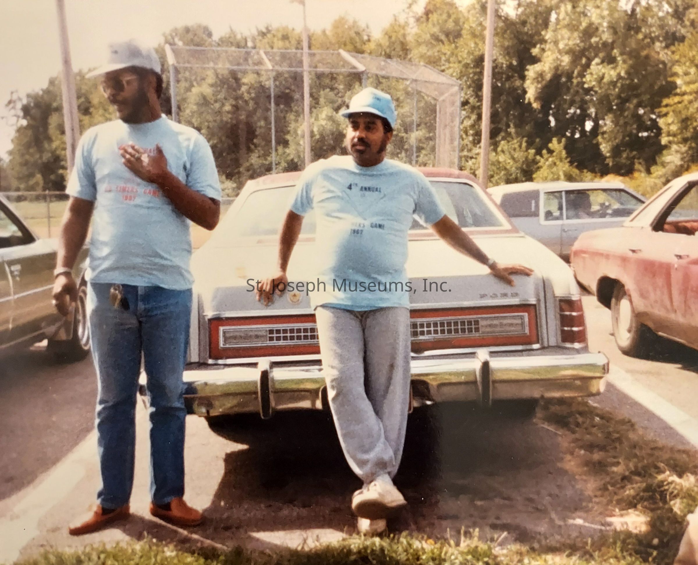 In the image, two men are standing near the back of an older car parked along a road, with large trees and a baseball backstop in the background. Both individuals are wearing light blue T-shirts with writing on them, and are also sporting blue caps. The man on the left, wearing a watch and keys dangling from his belt, is gesturing with his right hand while engaging in conversation. The man on the right is leaning casually against the car with his arm behind him and one foot crossed over the other, looking relaxed. The scene appears to be in a parking area, surrounded by several parked cars of similar vintage. The atmosphere seems relaxed and bright, suggesting it could be a sunny day, contributing to the warmth and camaraderie suggested by the scene.