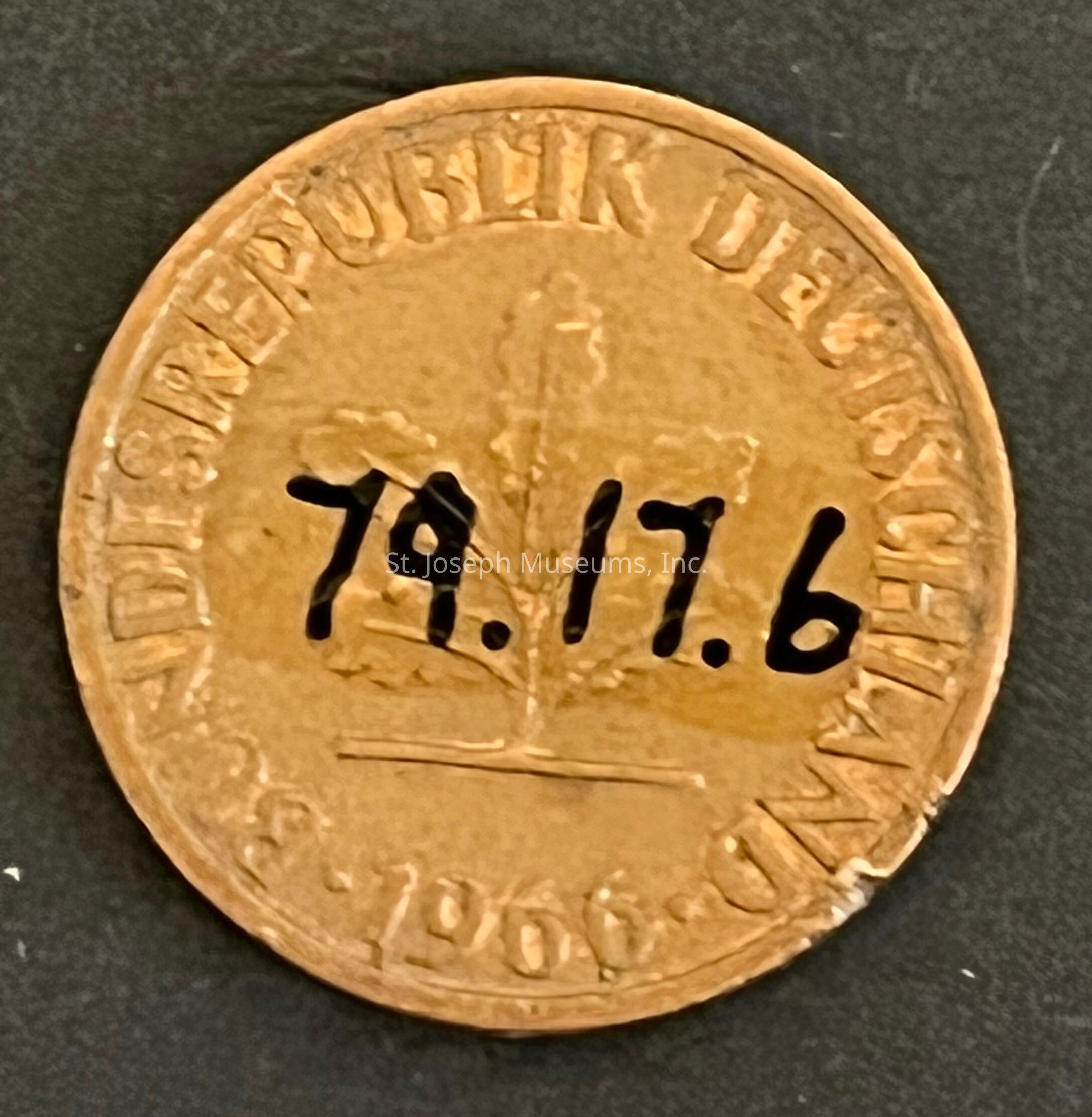 The image features a German coin, possibly from the Bundesrepublik Deutschland, with the date "1966" inscribed on it. The coin, likely made of a bronze-colored metal, has a central design of an oak leaf, which is a common symbol in German currency, representing strength and endurance. The upper inscription reads "BUNDESREPUBLIK DEUTSCHLAND," indicating the Federal Republic of Germany. In bold black marker, additional numbers "79.17.6" are written across the surface of the coin, possibly as inventory or identification markings. The dark background contrasts with the metallic sheen, highlighting the details and texture of the coin. The edges appear slightly worn, suggesting age or circulation. The coin’s surface has a patina, hinting at its vintage nature. The artistic design encompasses both functional monetary value and cultural symbolism, reflecting the historical period it represents. This image encapsulates both the utilitarian and artistic aspects of currency, merging financial history with numismatic interest.