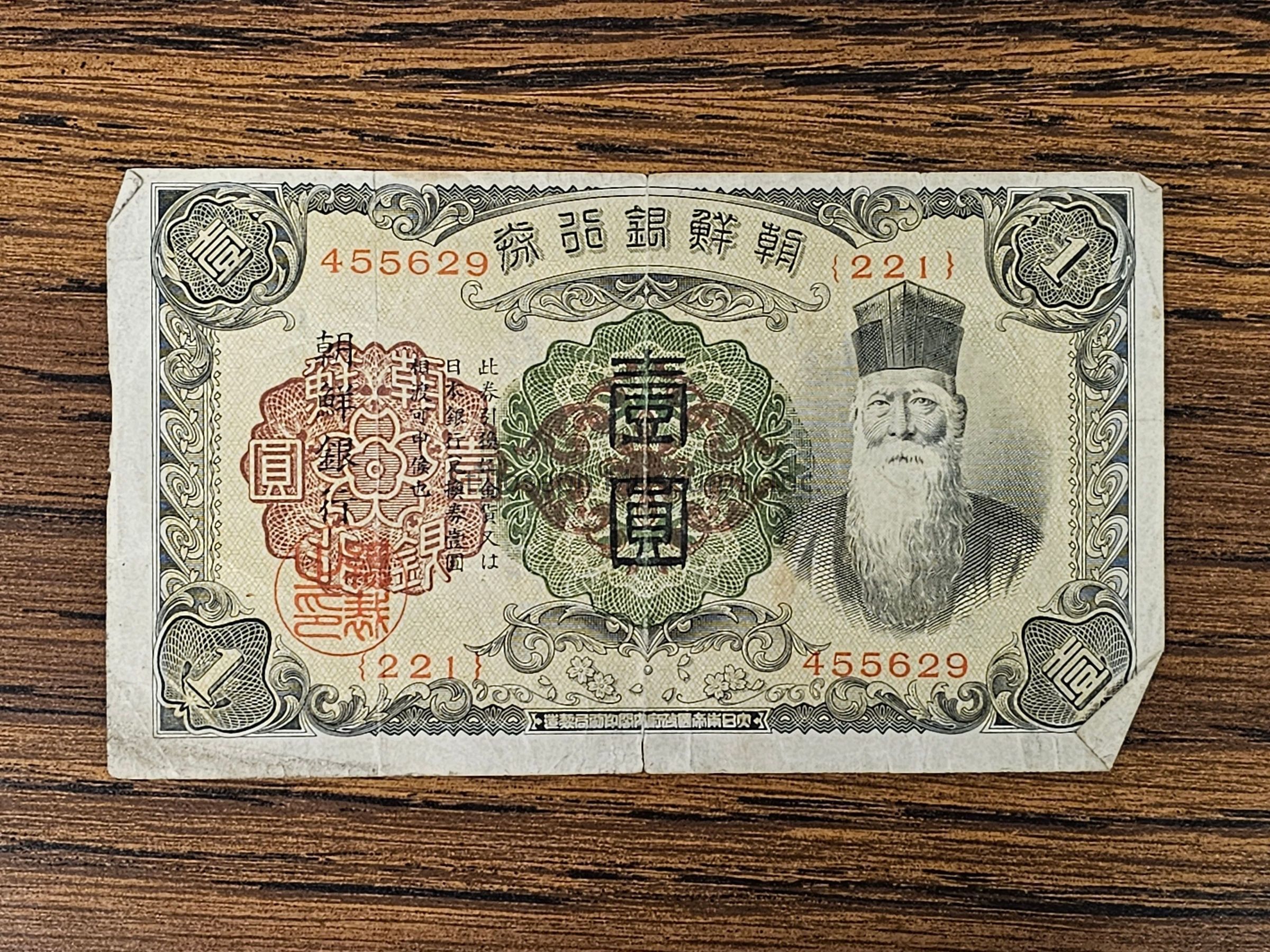 The image shows a vintage banknote from Korea with ornate designs and text in Korean characters. The top corners bear intricate patterns, while the top center displays serial numbers "455629" and "2211." A detailed circular design occupies the central part of the note, incorporating both red and green elements. To the right, there is a portrait of a bearded man wearing a traditional hat. The left side features red seals with more text.