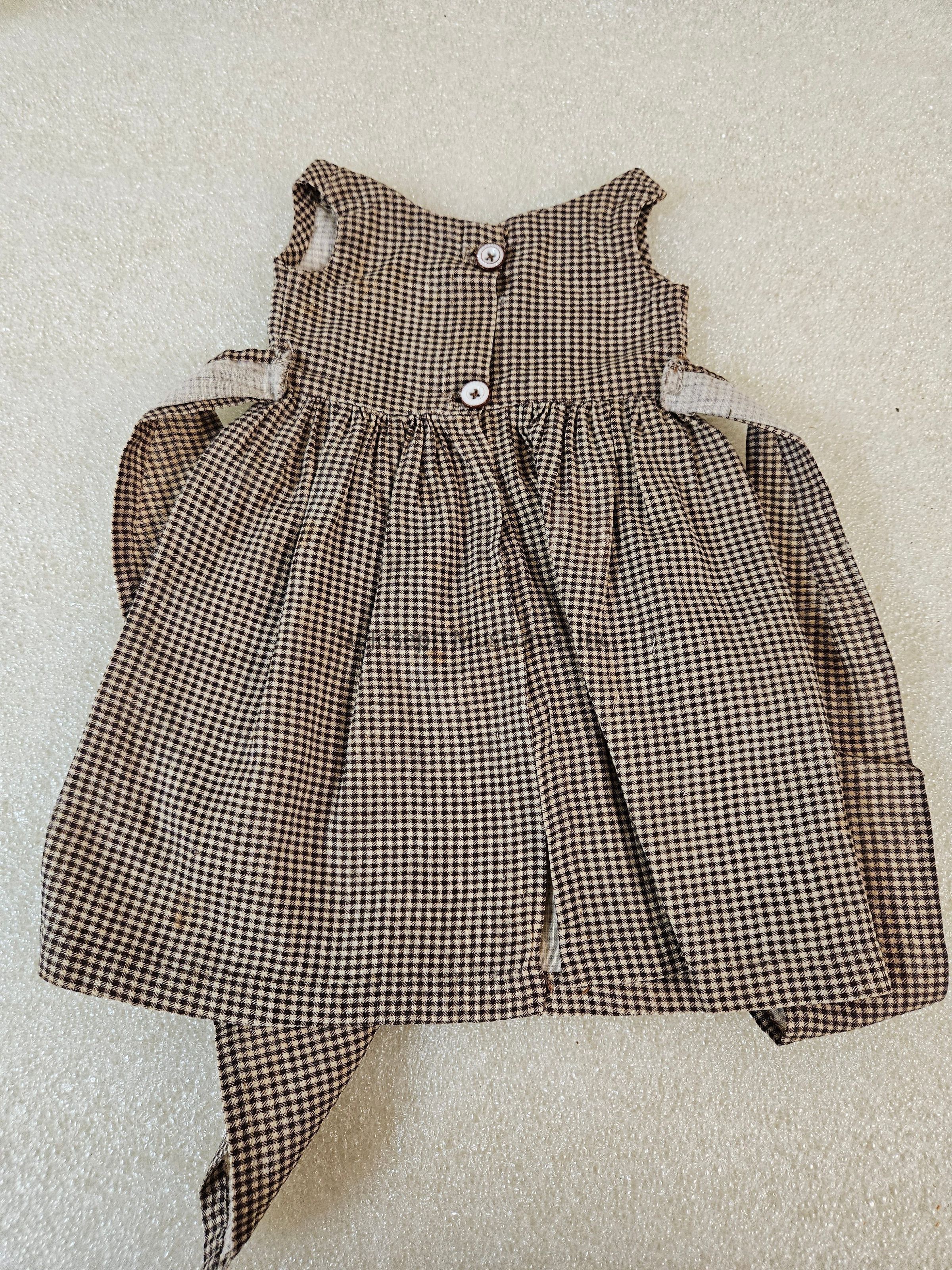 The image shows a children's dress with a classic checkered pattern in shades of brown and beige. It features a sleeveless design with wide armholes and a round neckline. The dress is buttoned down the back with two large, round buttons, adding both functional and decorative elements. The fabric appears to be lightweight and gathered at the waist, giving the skirt a full, flowing appearance. The dress is laid flat on a soft, textured, off-white surface, highlighting the craftsmanship and the pattern of the material. The overall style suggests a vintage or timeless aesthetic, suitable for various occasions, reflecting a sense of simplicity and charm.