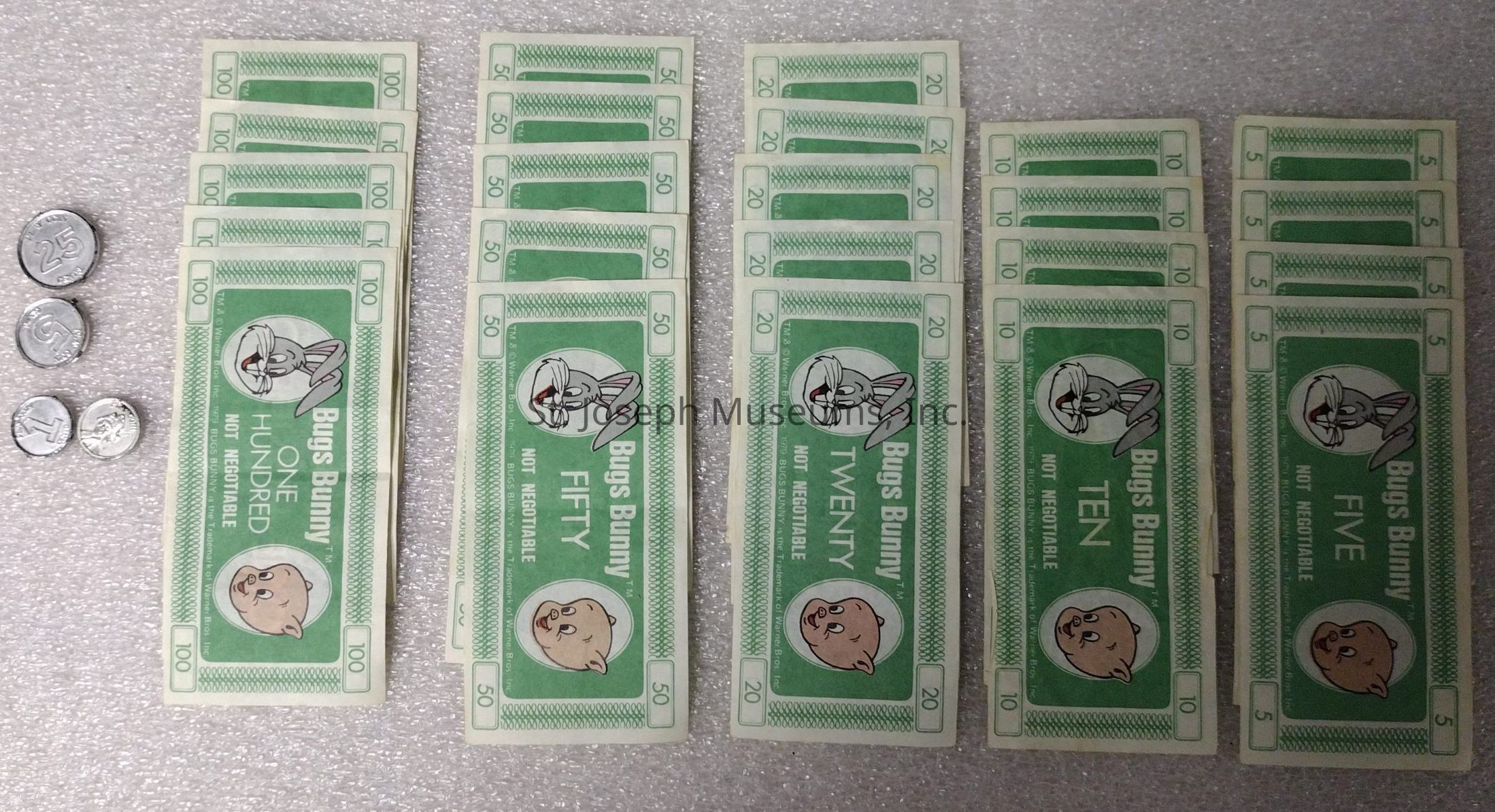 A: The image features a collection of themed play money bills and a few metallic coins on a textured surface. The bills display cartoon characters and are labeled as "Bugs Bunny" currency, indicating they are not real money. There are five columns of bills, each with a different denomination: 100, 50, 20, 10, and 5, depicted with varying shades of green. The denominations, which are clearly marked on each bill, feature the character Bugs Bunny's face prominently in a circular design on the left side, while a picture of another character is on the right. Additional text notes that they are "not negotiable." On the left side of the bills, four silver-colored coins are arranged below each other, hinting at their inclusion in a playful setup. The coins, different in size, stand out with their shiny metallic surface, contrasting with the matte finish of the paper bills. The overall arrangement and theme suggest a whimsical, cartoon-inspired aspect to the currency, making it
