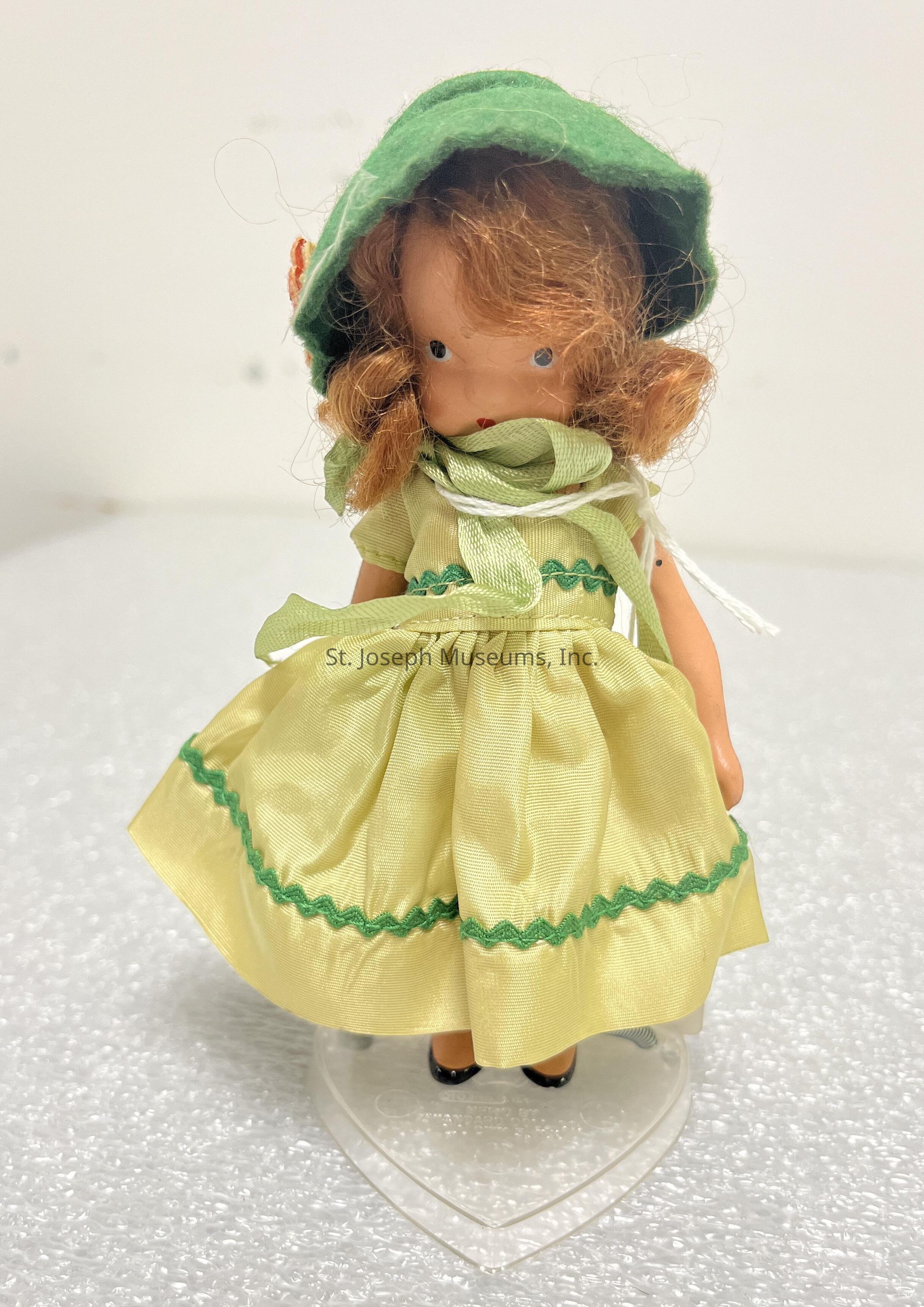 The image showcases a vintage doll with curly brown hair and a slightly tilted green felt bonnet, which matches the green sash around its neck. The doll is dressed in a light yellow dress adorned with green wavy trim detailing along the hem and bodice, giving it an old-fashioned charm. It stands upright on a clear heart-shaped stand that provides support while its small, black shoes peek out from beneath the flowing dress. The doll's painted facial features, including blue eyes and red lips, add a delicate, nostalgic touch.