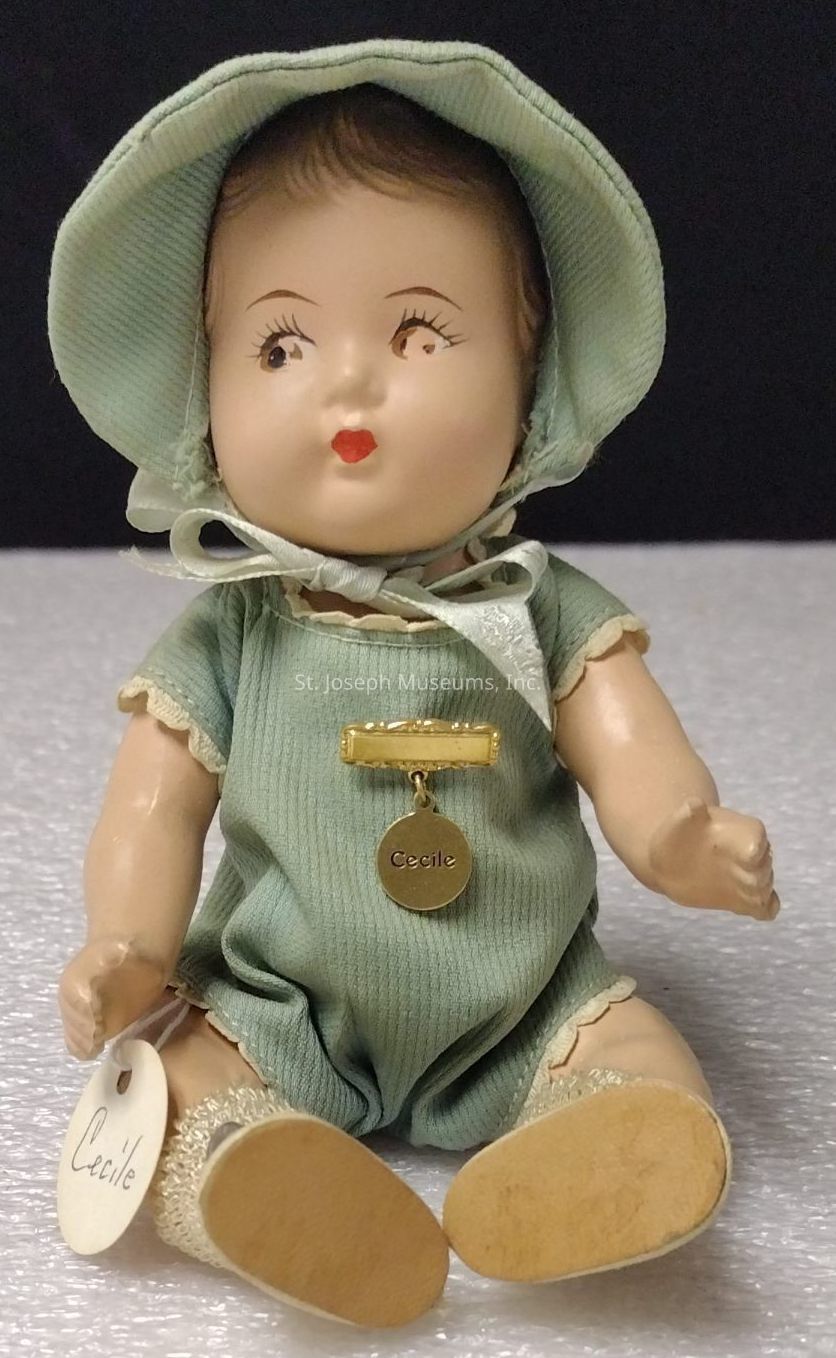 The image depicts a charming vintage doll clad in a light green outfit, complete with a matching bonnet. The doll is seated on a textured surface, and its attire, which features delicate lace trim, includes a short-sleeved romper that adds to its nostalgic appearance. A gold charm is pinned to the doll's chest, inscribed with the name "Cecile," suggesting a personalized touch or brand. The doll's facial features are meticulously crafted, with painted eyes and lips that convey an expression of innocent curiosity. Creased arms and legs reveal the doll's plush material, which indicates its role as both a collectible item and keepsake. Additionally, a tag with the name "Cecile" is attached to its wrist, complementing the gold charm and reinforcing the doll's identification or theme. The detailed craftsmanship of the doll, combined with its period-specific clothing and elegant accessories, evokes a sense of nostalgia, making it a treasured item for collectors and enthusiasts of vintage dolls.