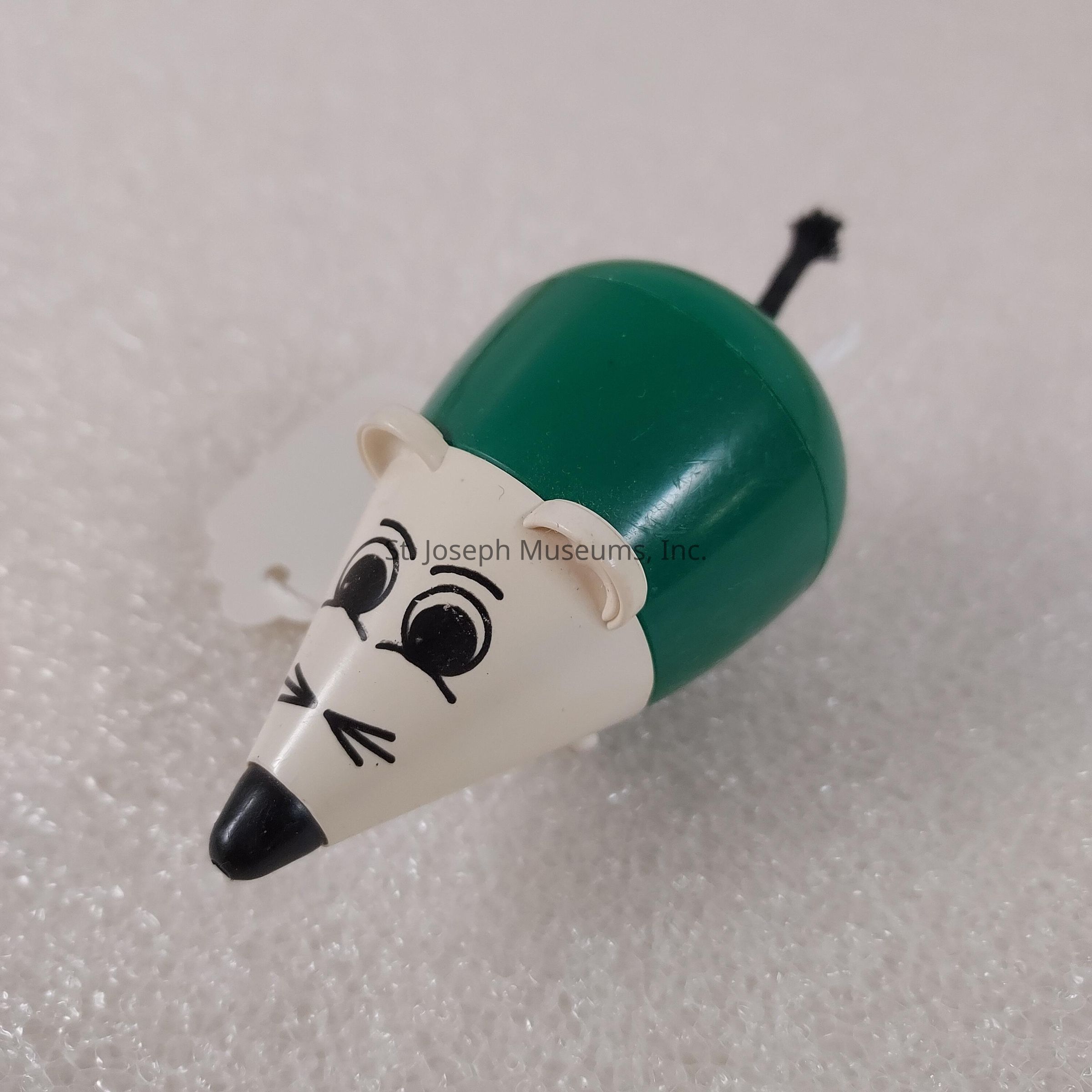 The image displays a small toy mouse with a body composed of a green, dome-shaped section and a white front. The mouse’s face is designed with simple black lines to depict features like eyes, whiskers, and a nose, while its ears are light beige in color. It has a small black tail extending from its green section. The toy rests on a textured, white, Styrofoam-like surface that provides a contrast to its colorful form. The overall design of the toy is whimsical and cartoonish, likely intended for entertainment or decorative purposes.