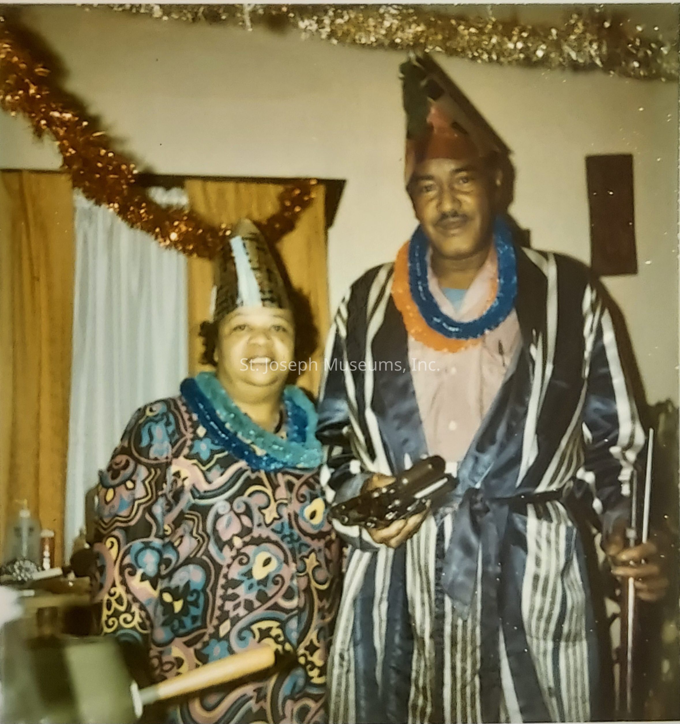 The image depicts a cheerful couple standing indoors, adorned in festive attire for a celebratory occasion. Both individuals wear colorful party hats and leis, indicative of a joyous event or party, likely during the holiday season. The woman is dressed in a vibrant, patterned outfit while the man sports a striped robe over his clothes, complementing the lively atmosphere. Behind them, shimmering garlands hang across the room, enhancing the festive theme. The expressions on their faces radiate happiness and warmth, suggesting they are enjoying this special moment together.