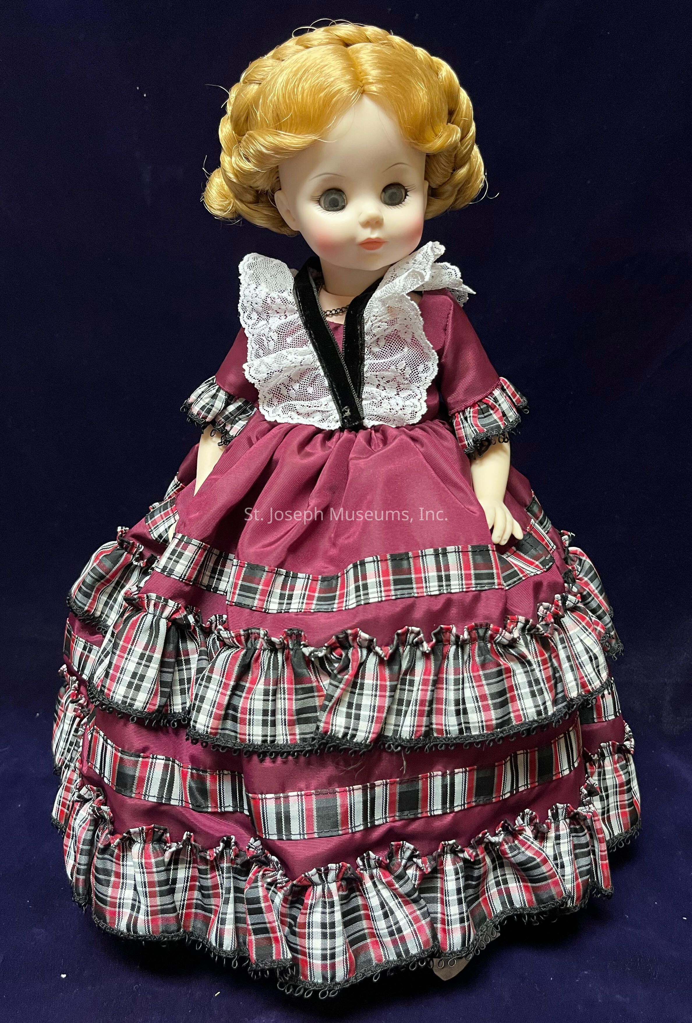 The image features a porcelain doll of Betty Taylor Bliss, the daughter of Zachary Taylor. The doll has curly blonde hair styled in an intricate updo. The doll is dressed in an elaborate burgundy gown adorned with layers of ruffled plaid fabric in red, black, and white. Additional details include a delicate white lace collar and black velvet trim accentuating the dress. The doll's facial features are delicately painted, with large, bright eyes and subtle blush on its cheeks.