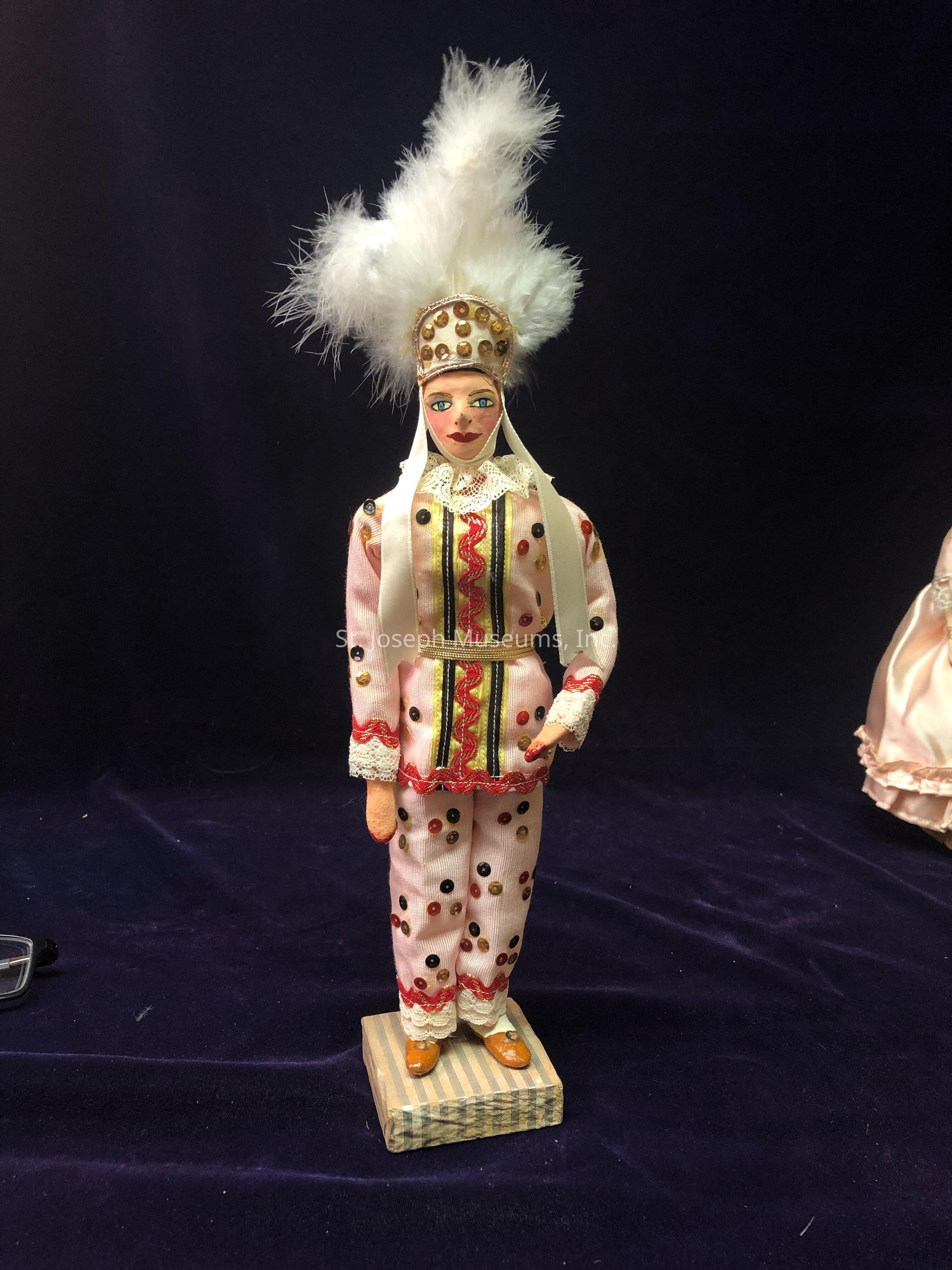 The image depicts a handcrafted doll dressed in an ornate and colorful costume. The doll is adorned with a bright outfit featuring vibrant patterns, including red and gold embellishments on a light-colored base. It wears a distinctive headdress decorated with golden elements and topped with large white feathers, enhancing its regal appearance. Standing on a striped pedestal, the doll showcases intricate detailing, such as lace trim at the cuffs and collar, alongside embellishments and sequins throughout the attire. The background is a dark, rich fabric, which contrasts with the doll’s vivid attire, highlighting its vibrant colors and craftsmanship. The overall presentation suggests a theatrical or ceremonial theme, possibly representing a traditional costume or cultural attire from a specific region or event. The expression on the doll's face is serene and stylized, with delicately painted features that add to its character and charm.