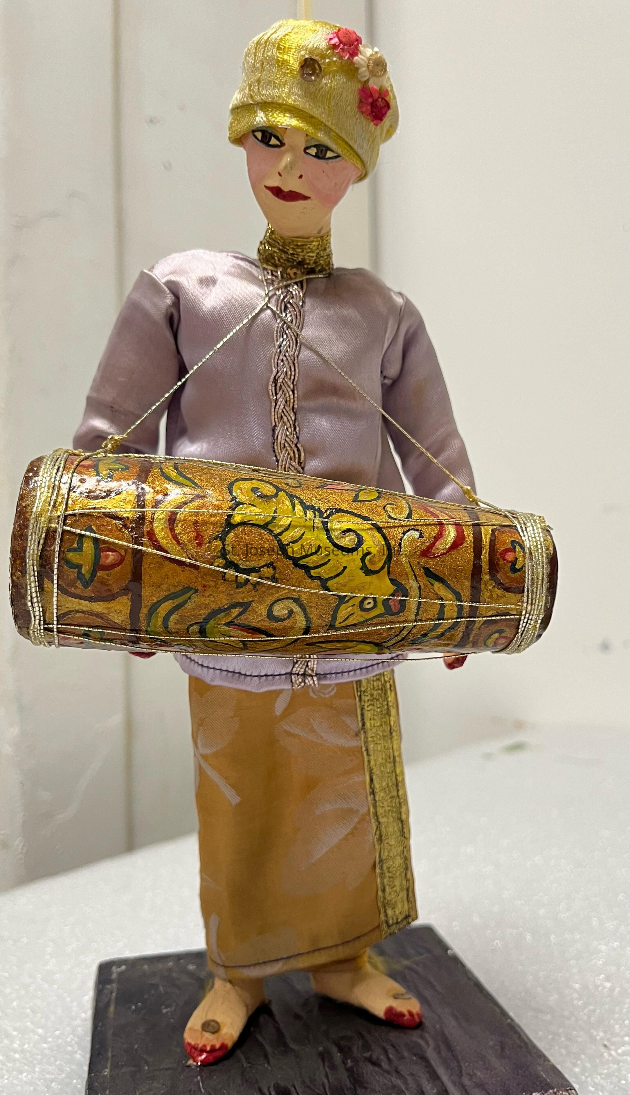 The image shows a handcrafted doll dressed in a traditional attire, featuring a soft lavender top with intricate silver detailing down the front and a mustard-colored long skirt. The doll wears a matching gold hat adorned with small, delicate fabric flowers in pink and beige tones. Its face is carefully painted with fine details, including expressive eyes and red lips. Across the doll’s shoulder, it carries a richly decorated drum with yellow and orange patterns and floral designs, secured with thin gold cords. The doll stands on a simple dark wooden base, adding elegance.