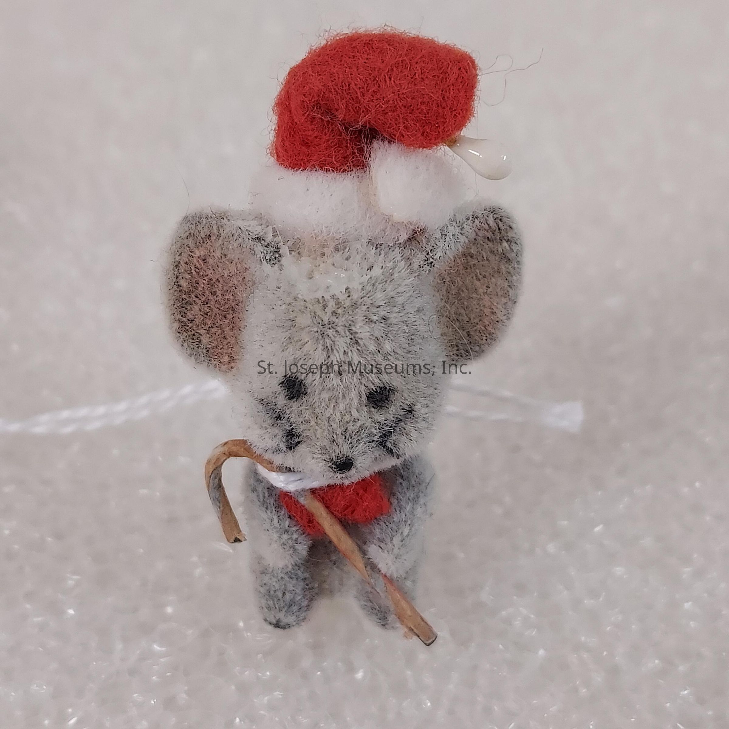 The image depicts a small, handcrafted mouse ornament, featuring a gray and fuzzy texture with large, rounded ears and a simple face. This charming mouse is adorned with a tiny red Santa hat that adds a festive touch, enhancing its overall cuteness and holiday spirit. Held in its diminutive paws is a miniature candy cane crafted from what appears to be a thin piece of material, likely intended to mimic wood or plastic, providing it with a quaint, rustic look. The mouse wears a small red scarf around its neck, adding to the warmth and coziness of its appearance, while suggesting it is well-prepared for the chilly Christmas season. The background features a textured, possibly foamed white surface that gives the image a soft, wintry feel and enhances the holiday theme by creating a subtle contrast with the predominantly gray and red hues of the mouse ornament. The mouse's endearing expression and posture convey an innocent and friendly demeanor, making it a delightful addition to any holiday decoration collection.