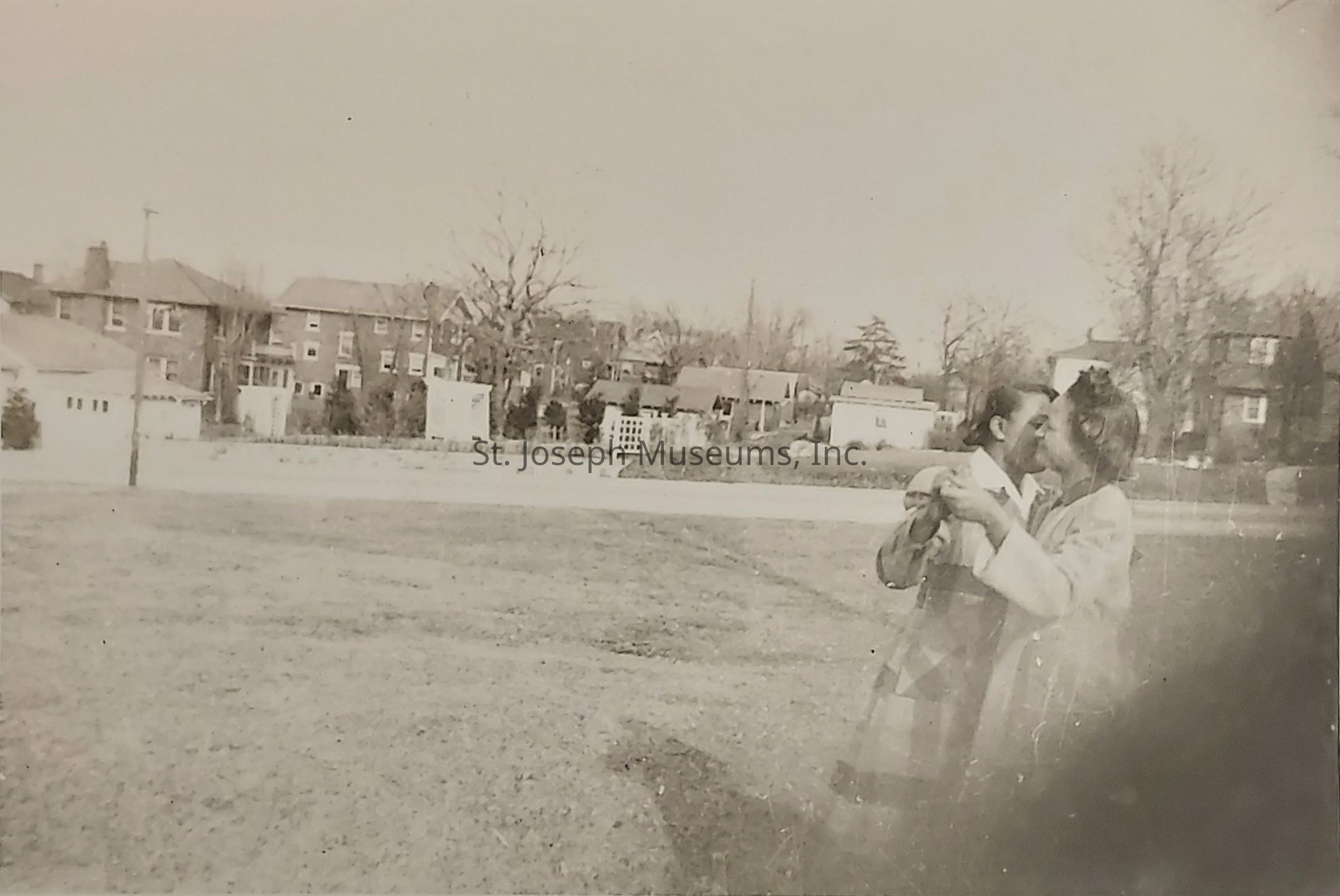 In a nostalgic black and white photograph, two individuals share a tender kiss in the foreground of an expansive grass field, surrounded by a quiet residential neighborhood in the background, with a line of suburban houses characterized by their distinct brick facades, chimneys, and quaint gardens, while bare deciduous trees dot the landscape, casting subtle shadows on the ground, suggesting a season of early spring or late autumn; the distant horizon meets a pale, almost overexposed sky, creating a serene and timeless atmosphere that captures a moment of intimacy and affection amidst the simplicity of everyday life, highlighting the enduring human connection that transcends the passage of time and the mundane backdrop of suburban routine.