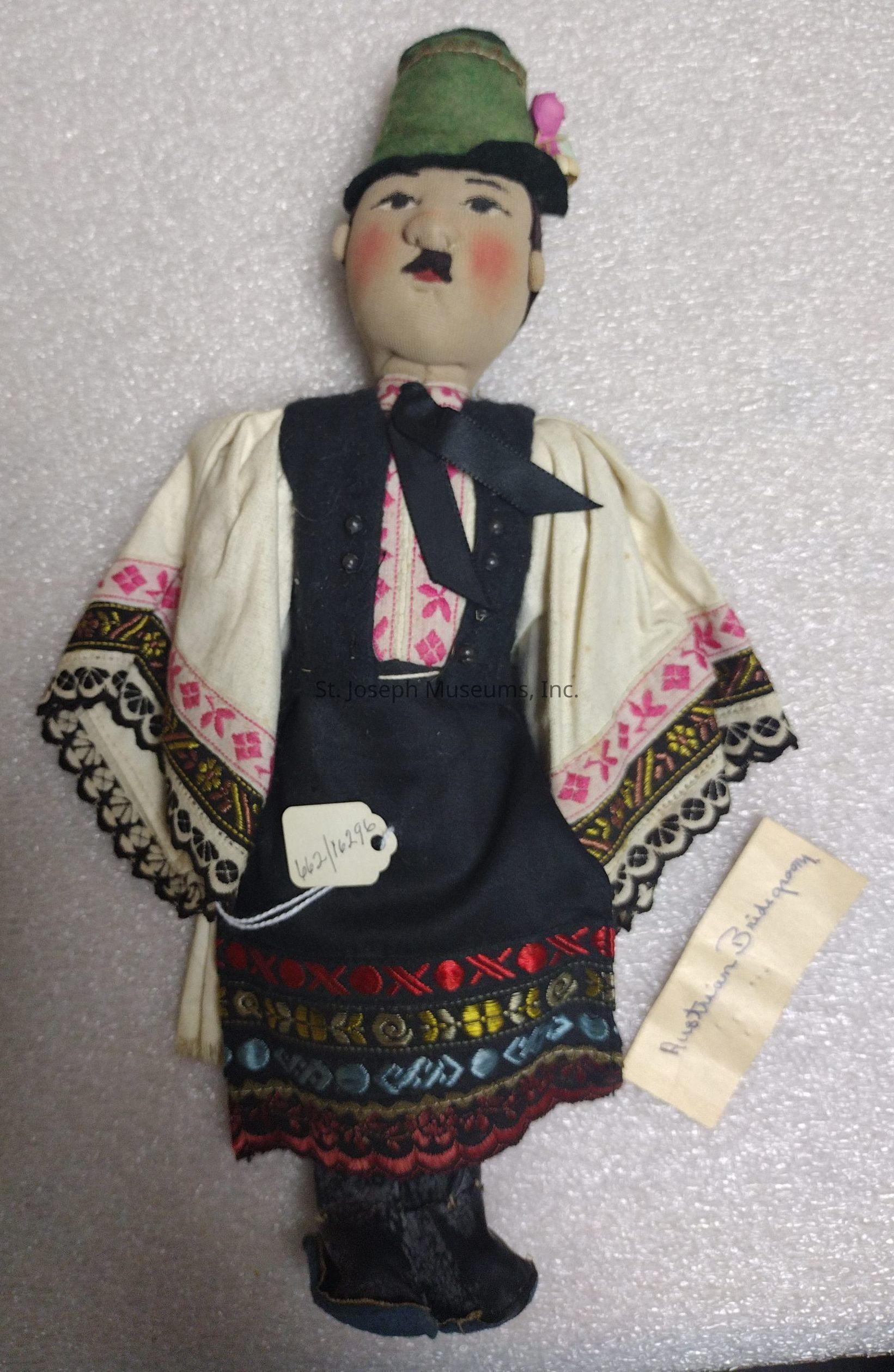 The image depicts a handcrafted doll adorned in traditional attire, featuring an intricate and colorful embroidered design on its garments. The doll is dressed in a white blouse with pink and black geometric patterns, which are enhanced by detailed embroidery along the sleeves. It wears a dark vest with small buttons and a black apron with a decorative tag attached. The skirt displays a vibrant array of colors including red, blue, and gold, showcasing an elaborate design. The doll's face is painted with distinct features, including rosy cheeks and expressive eyes, topped with a green felt hat embellished with small flowers. The overall craftsmanship and attention to detail suggest cultural significance, evident from the label that reads "Austrian Brautjunge," hinting at possible origins.