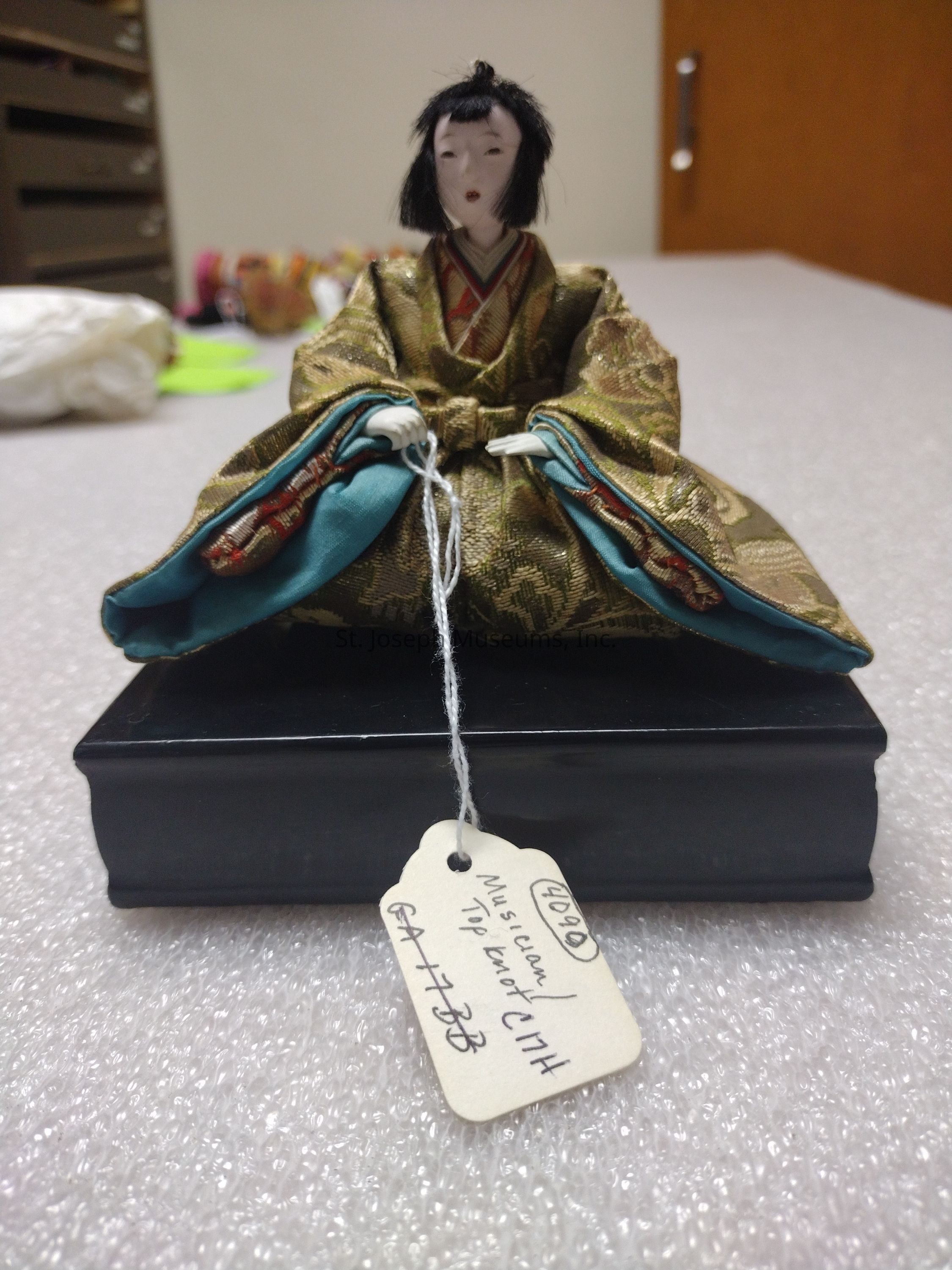The image depicts a small, intricately crafted figurine of a musician, sitting cross-legged atop a black, rectangular base. This figurine features a serene expression and wears a traditional, elaborate garment adorned in a pattern of gold and green, with accents of turquoise lining, which drapes over and around the body. The figure's head is adorned with short, black hair styled in a neat manner. In front of the figurine, a small, handwritten tag is attached with a string, displaying text including the number "40990" and the words "Musician" and "Top Knot," possibly indicating the item’s catalog number and description. The background of the image reveals a slightly blurred room setting with various objects in soft focus, suggesting the environment may be part of a storage or archival space. The composition emphasizes the delicacy and detail of the figurine, showcasing its artistic and cultural significance.