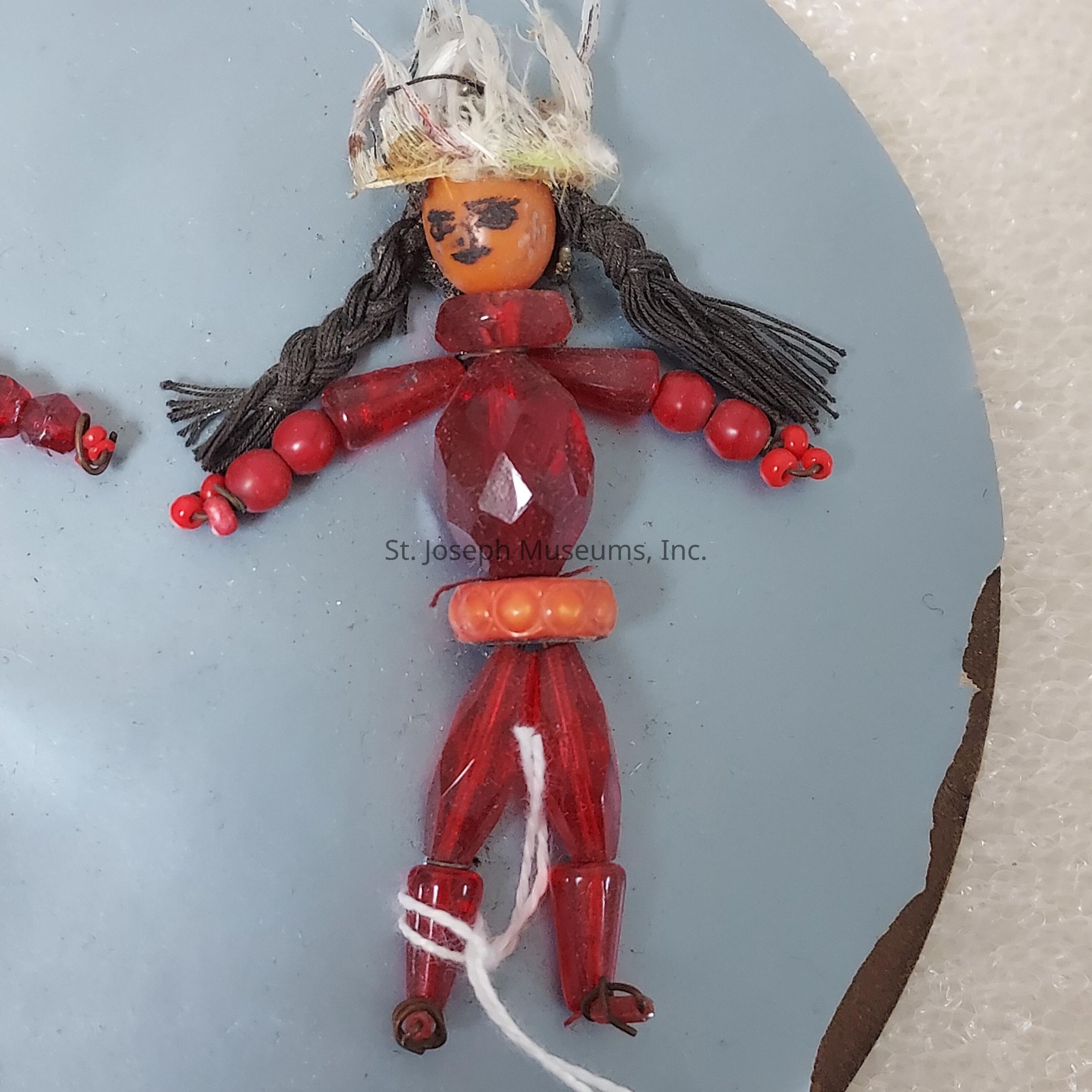 The image shows a handmade figure crafted from various beads, featuring a small doll-like design. The head is made from a wooden bead with a painted face, showcasing an expression with two dot eyes and a small mouth. It is adorned with a white feather-like material on top, resembling hair. The body consists of red faceted beads forming a torso, with smaller round beads creating the arms and legs. The figure is tied together with white string, adding a touch of intricacy to the construction. The doll appears to be mounted on a plain, light blue background, giving it a distinct, standout appearance. The use of red beads contrasts against the serene blue backdrop, highlighting the doll. It possesses braided black strands resembling hair extensions on either side of the head. Around the waist, an orange bead serves as a decorative element, adding visual interest. There are tiny, additional red beads on the arms, resembling hands, enhancing its detailed handcrafted quality.